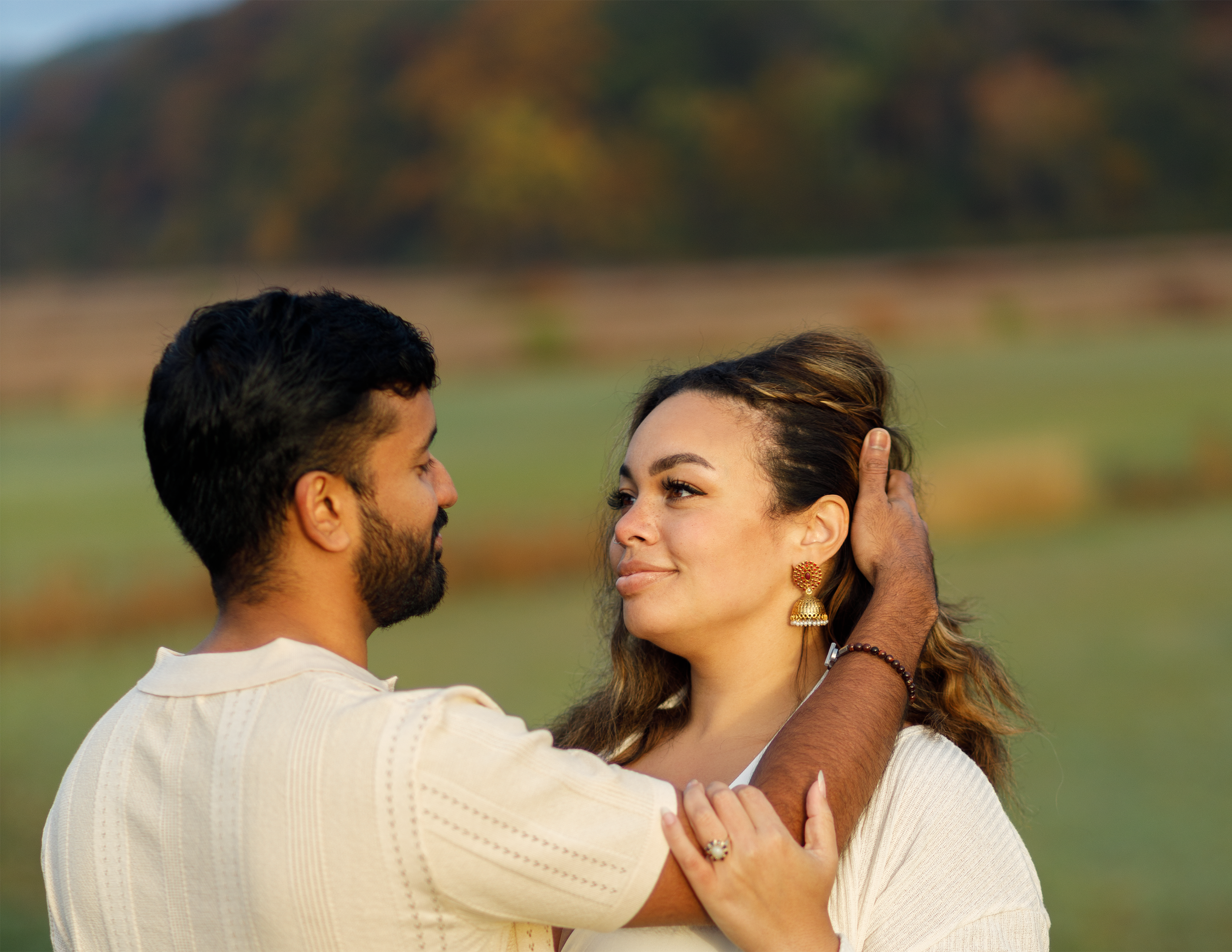 A man gently holding a woman's face with one hand while looking at her affectionately in a park with greenery and trees in the background.