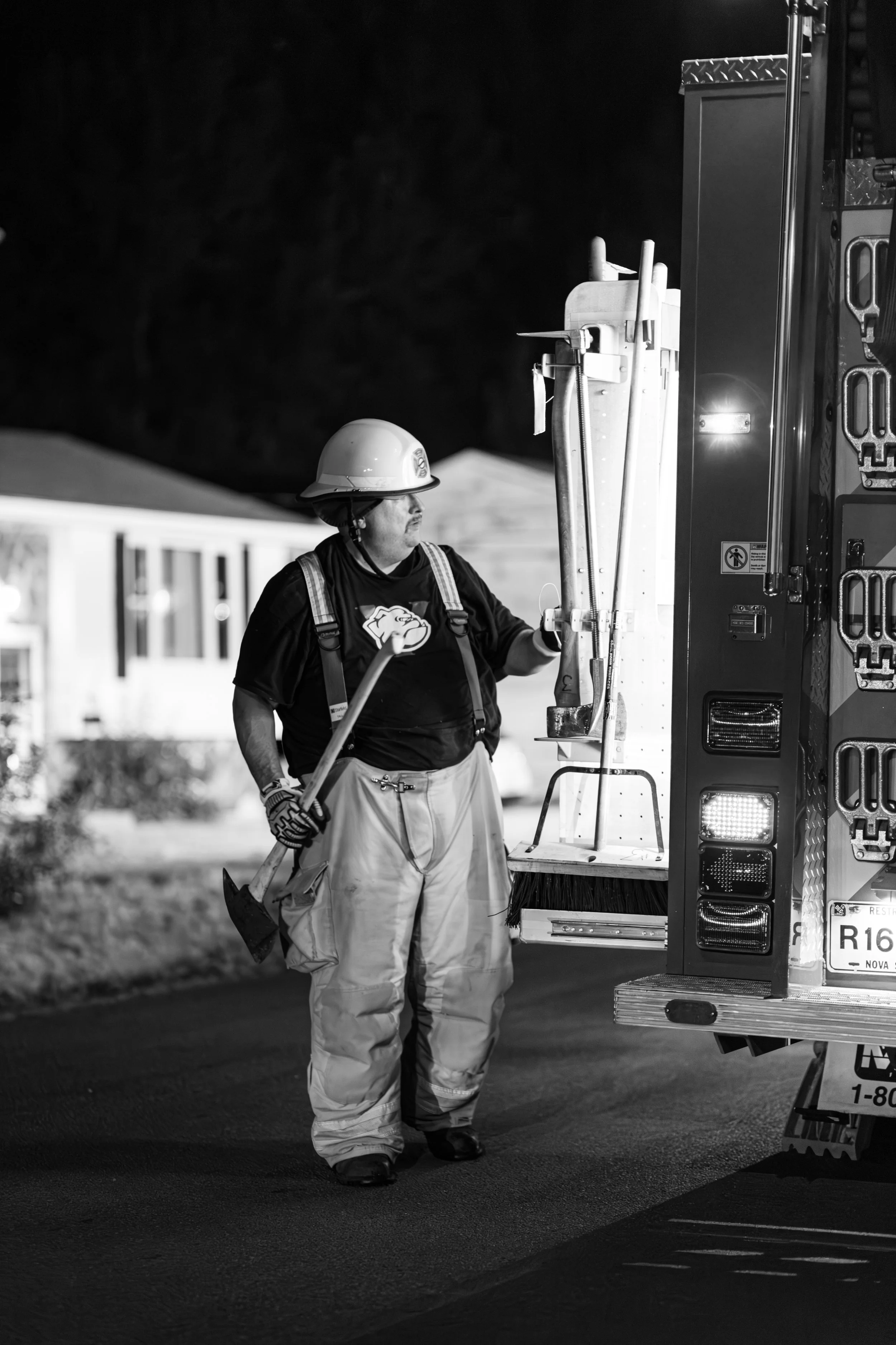 Firefighter wearing a helmet and protective clothing standing by a fire truck at night, holding an axe.