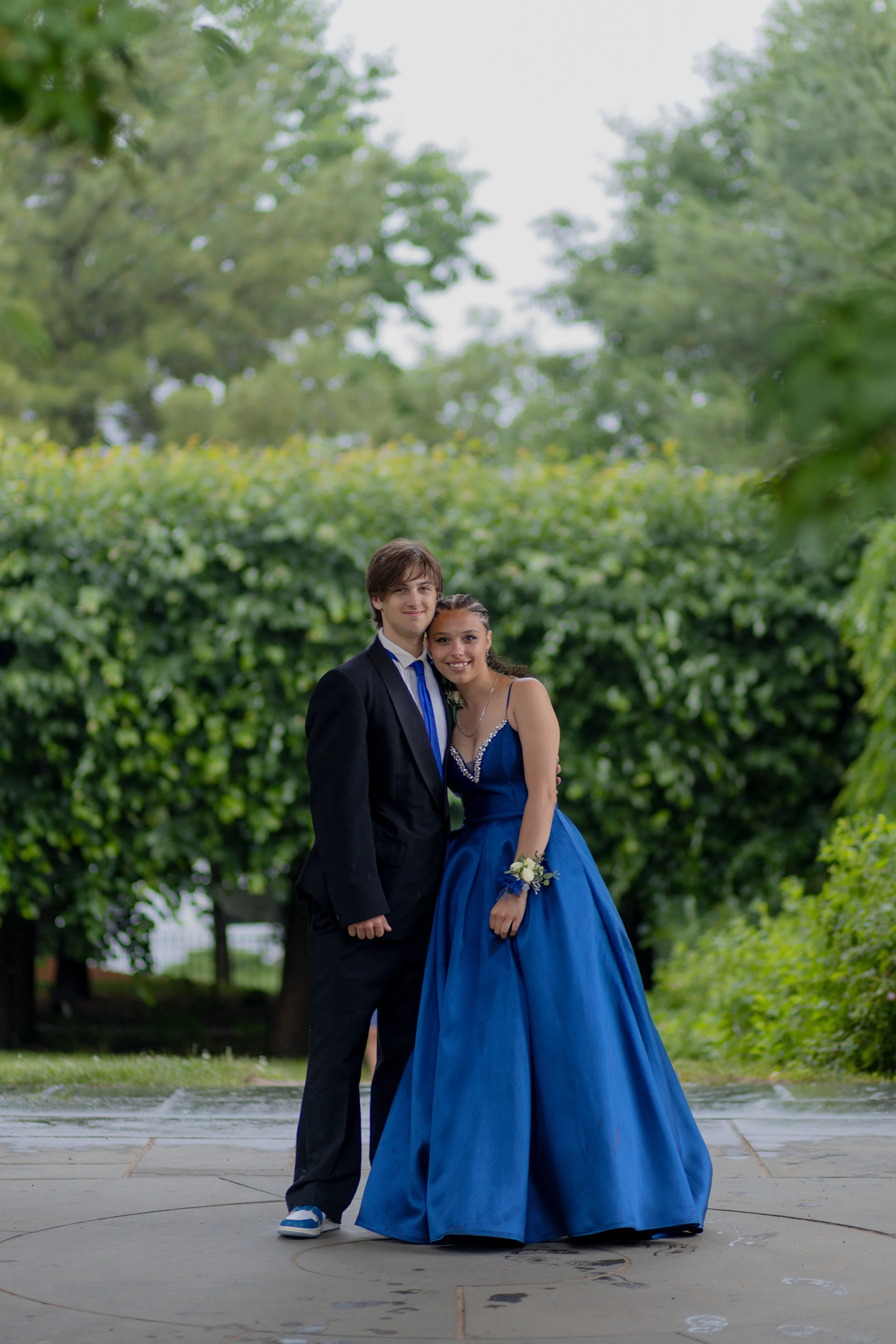 Teenagers dressed in formal attire, standing on a paved walkway in a park with green trees in the background. The girl is wearing a blue gown and corsage, and the boy is in a black suit and blue tie.