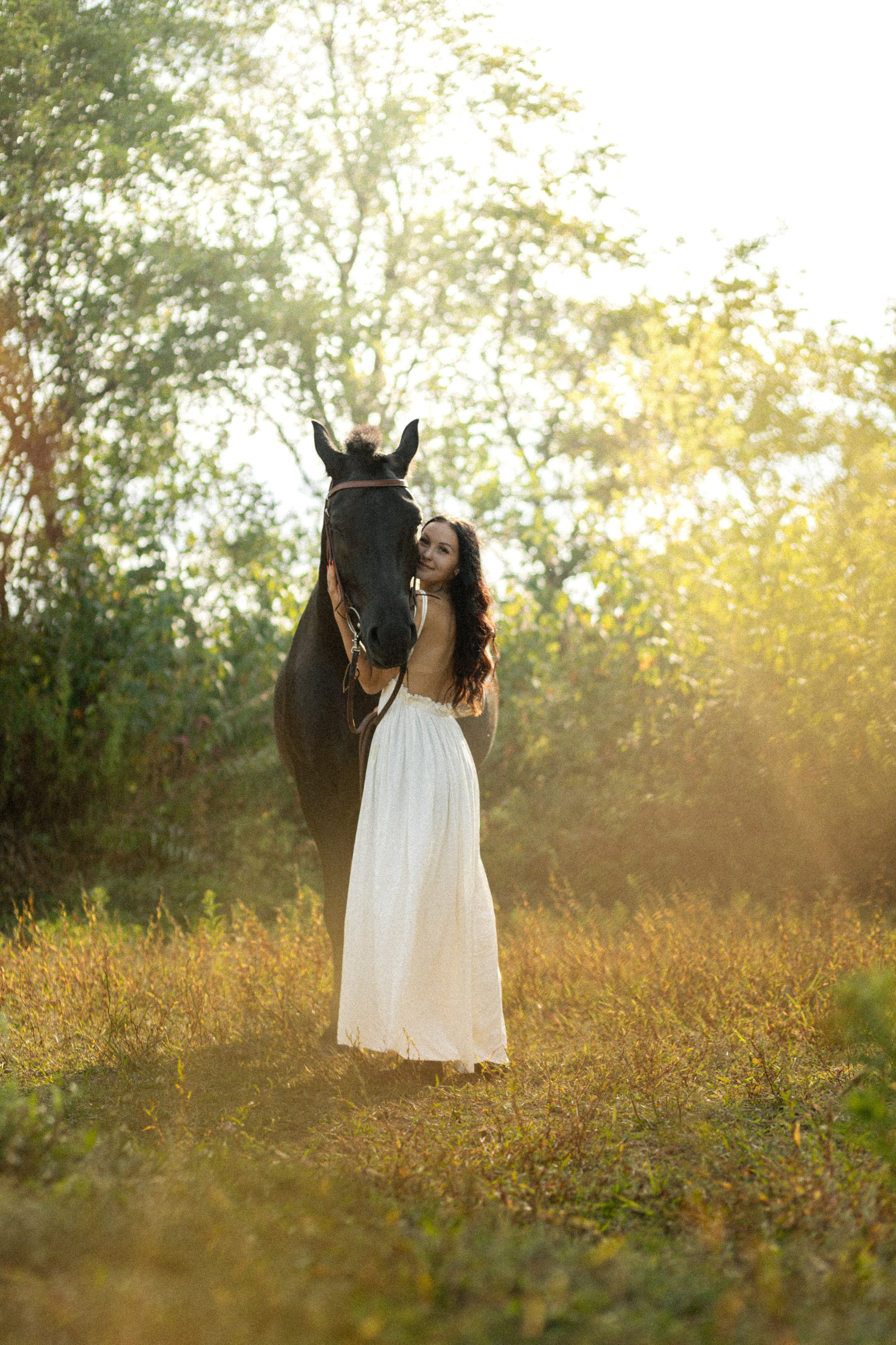A woman in a white dress standing next to a black horse in a sunlit outdoor setting with trees in the background.