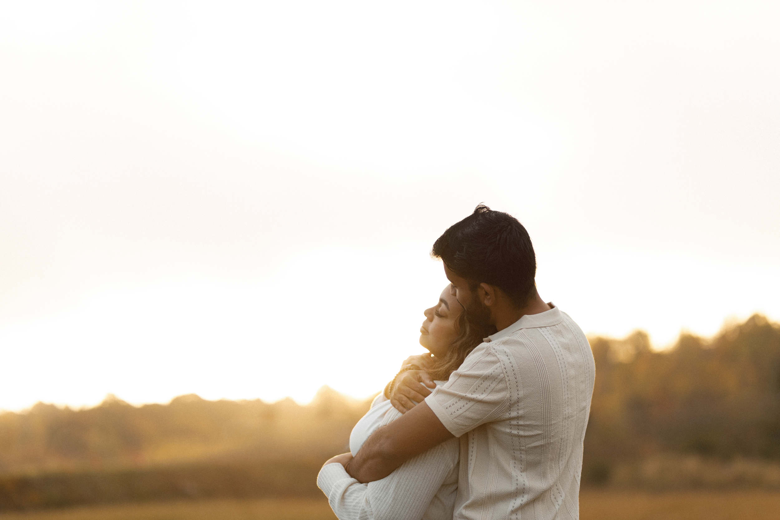 A couple embracing outdoors during sunset, with the man holding the woman close and the woman resting her head on his shoulder, against a blurred natural background.