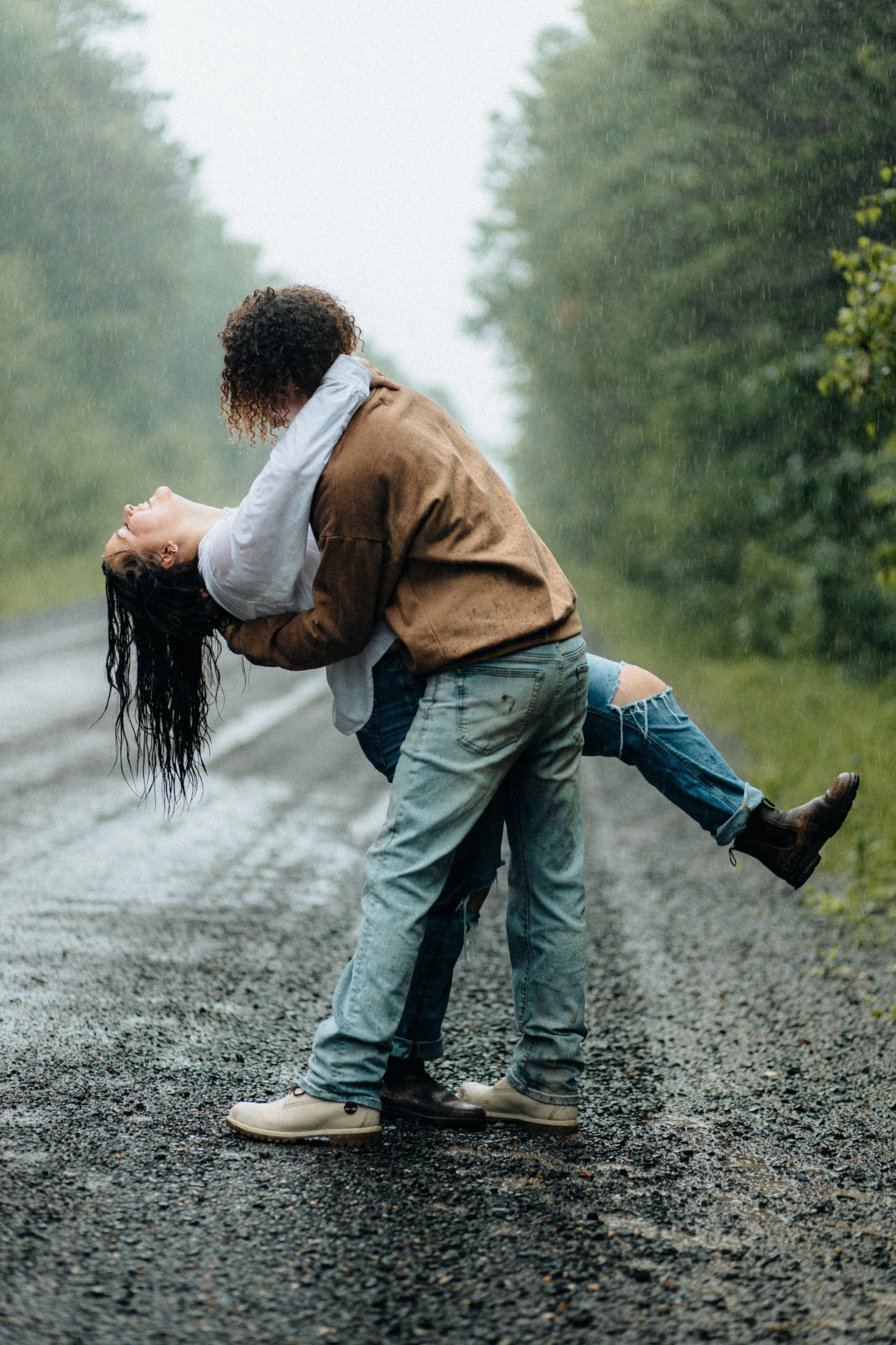 Two women are dancing and embracing on a rainy, gravel road surrounded by greenery, with one woman wearing a brown jacket and the other in ripped jeans and boots.