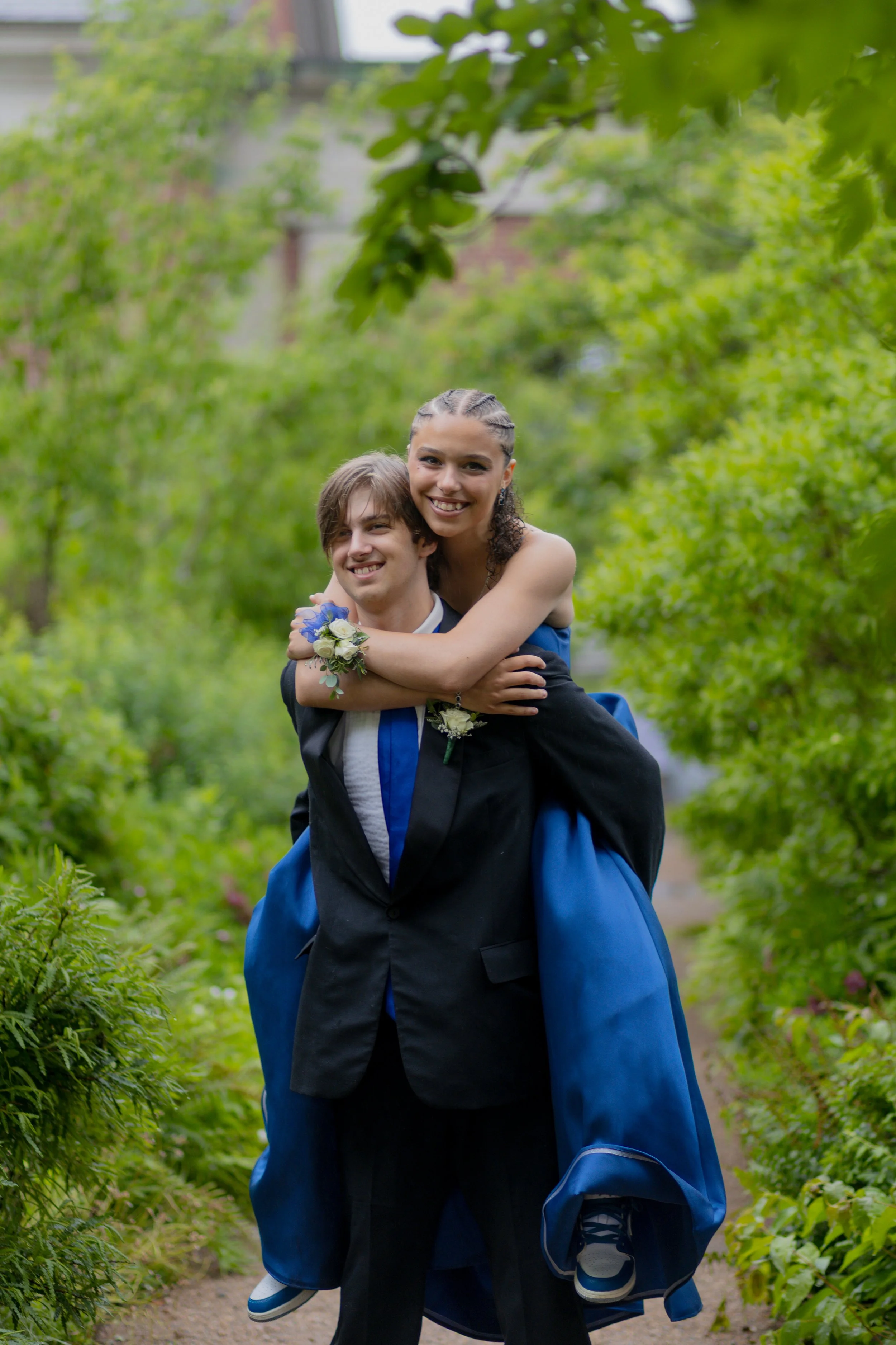 A young man in a black graduation gown carrying a young woman in a blue gown on his back outdoors on a green pathway, both smiling.