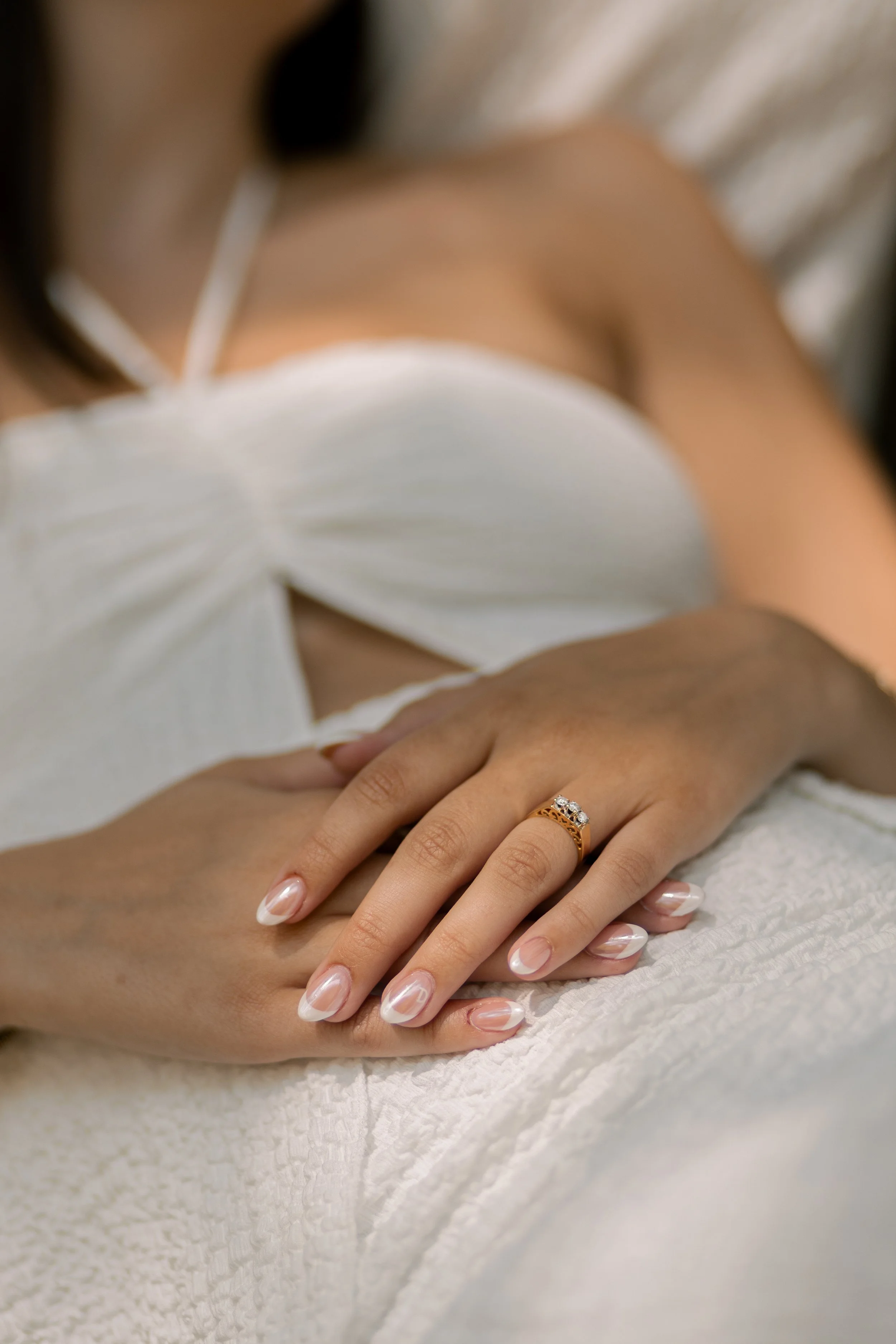 Close-up of a woman's hand resting on a white textured fabric, displaying a gold ring with diamonds on her ring finger. The woman is wearing a white outfit with a sweetheart neckline.