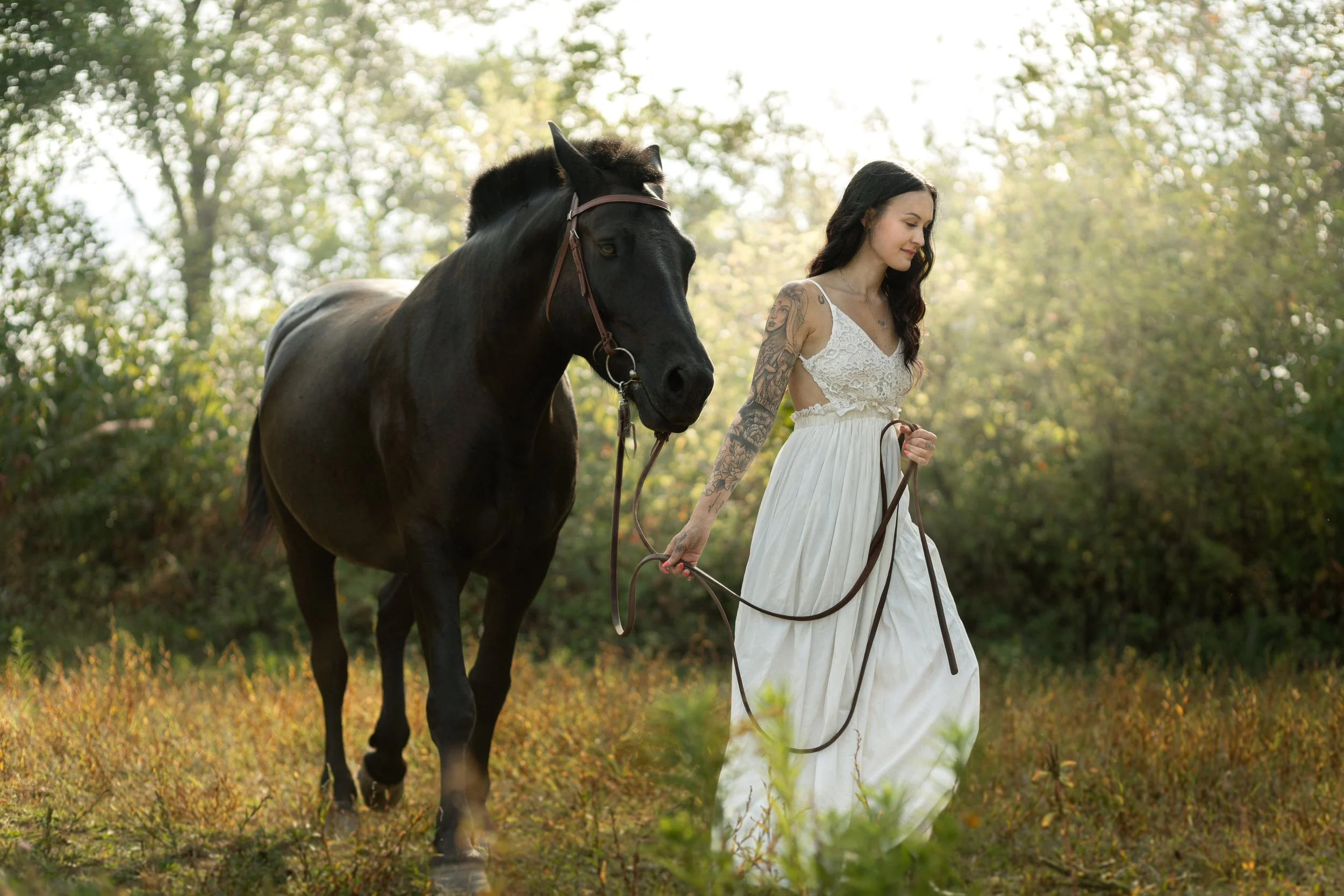 A woman with long dark hair, wearing a white lace dress with cutouts, holding a brown leash attached to a black horse, walking through a sunlit, wooded area with green foliage.