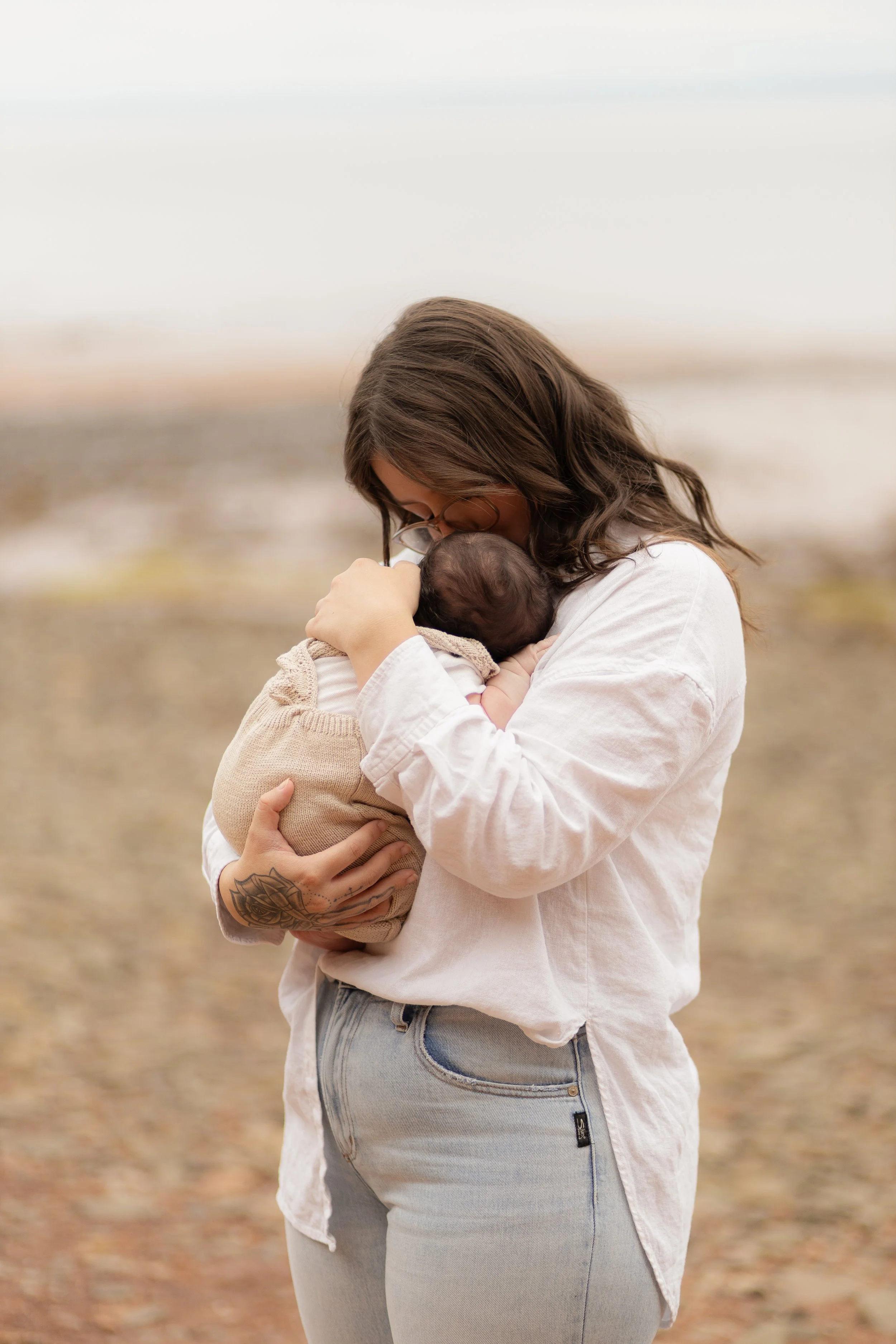 A woman holding and gently kissing a newborn baby at the beach.