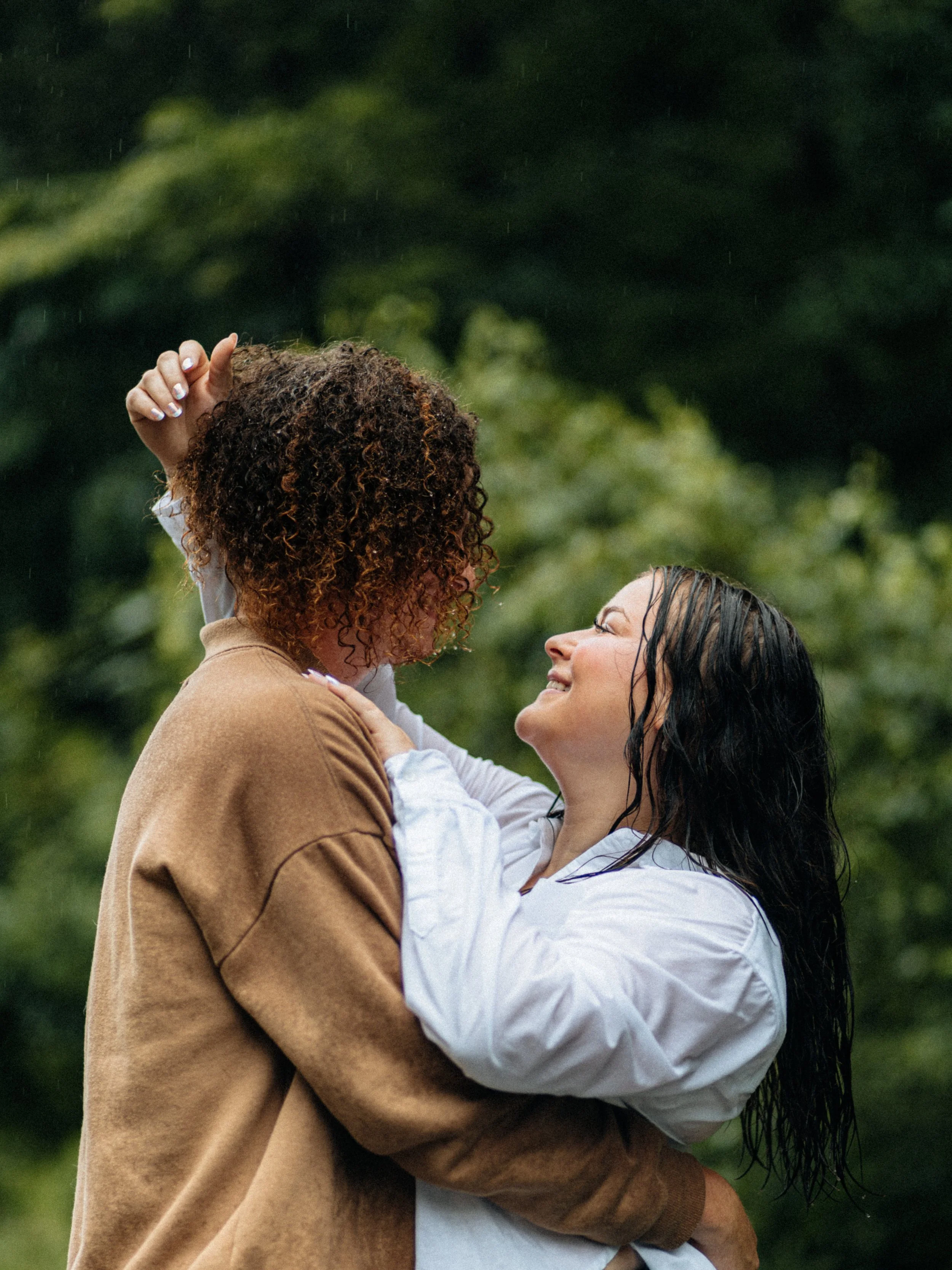 Two women, one with curly hair and the other with wet straight hair, embrace outdoors in a green, forested area, smiling at each other.