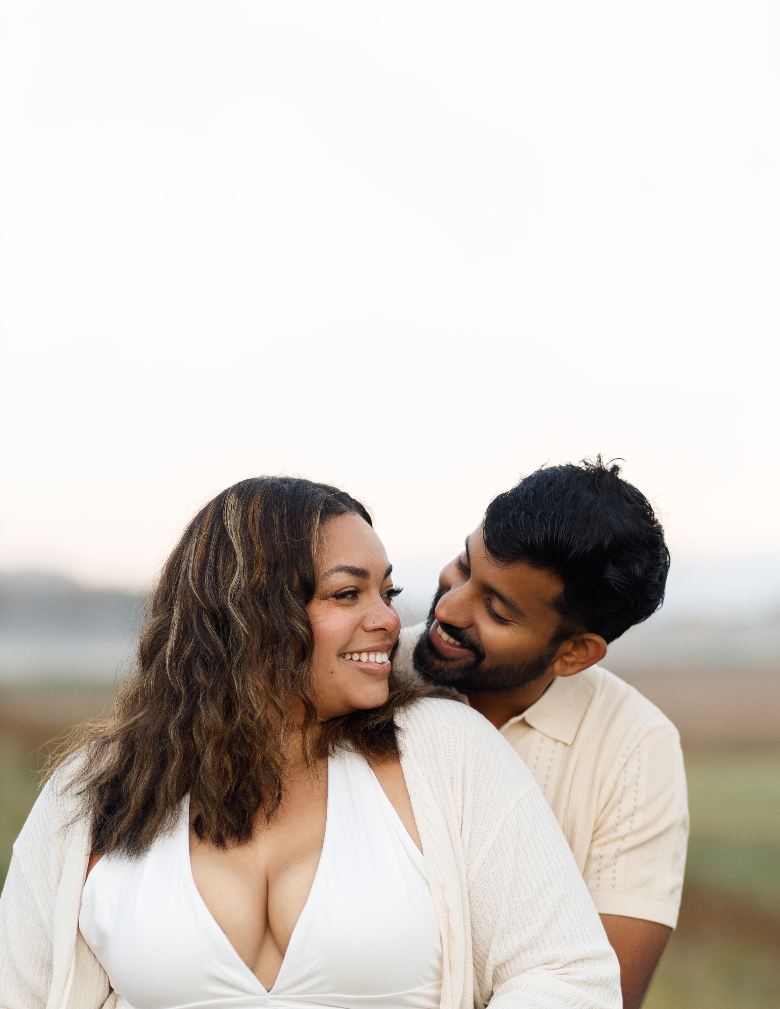 A smiling woman with curly hair and a man with dark hair and a beard sharing an affectionate moment outdoors.