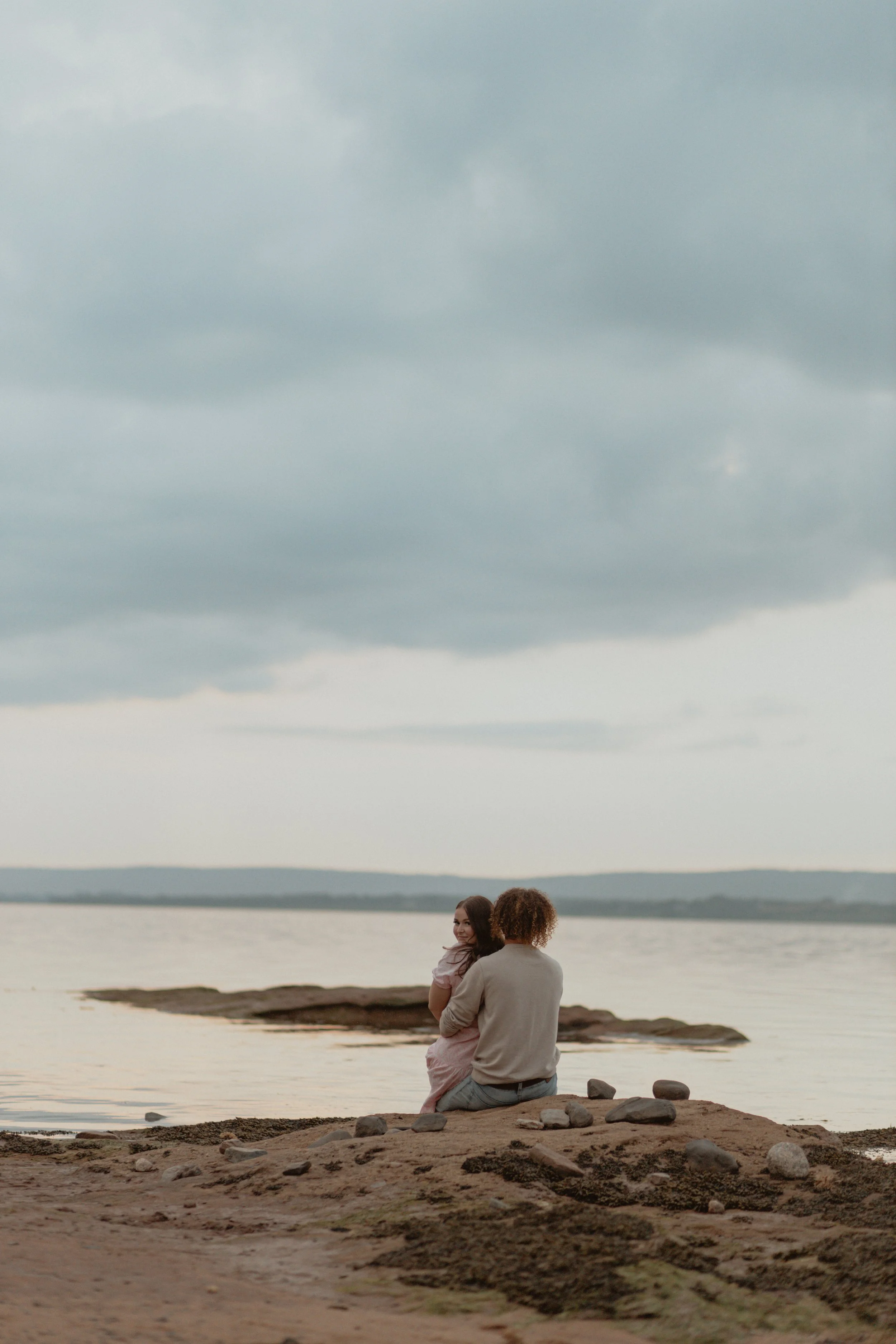 A person sitting on a rock on the sandy shore, holding a child, with a calm body of water and overcast sky in the background.