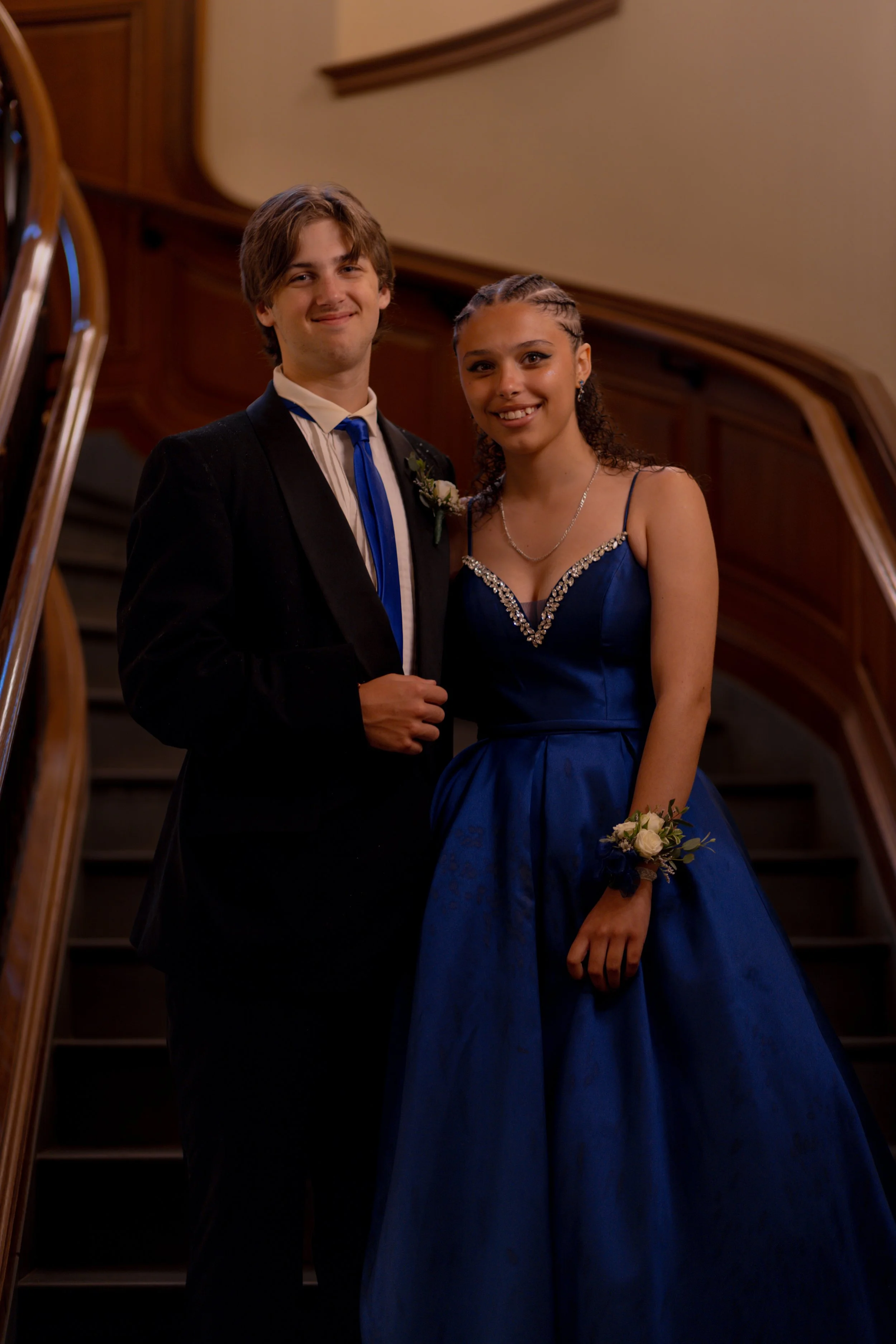 A young couple in formal attire standing on a staircase, smiling at the camera. The man wears a black tuxedo with a blue tie and a boutonniere, and the woman wears a blue gown with embellishments and a wrist corsage.