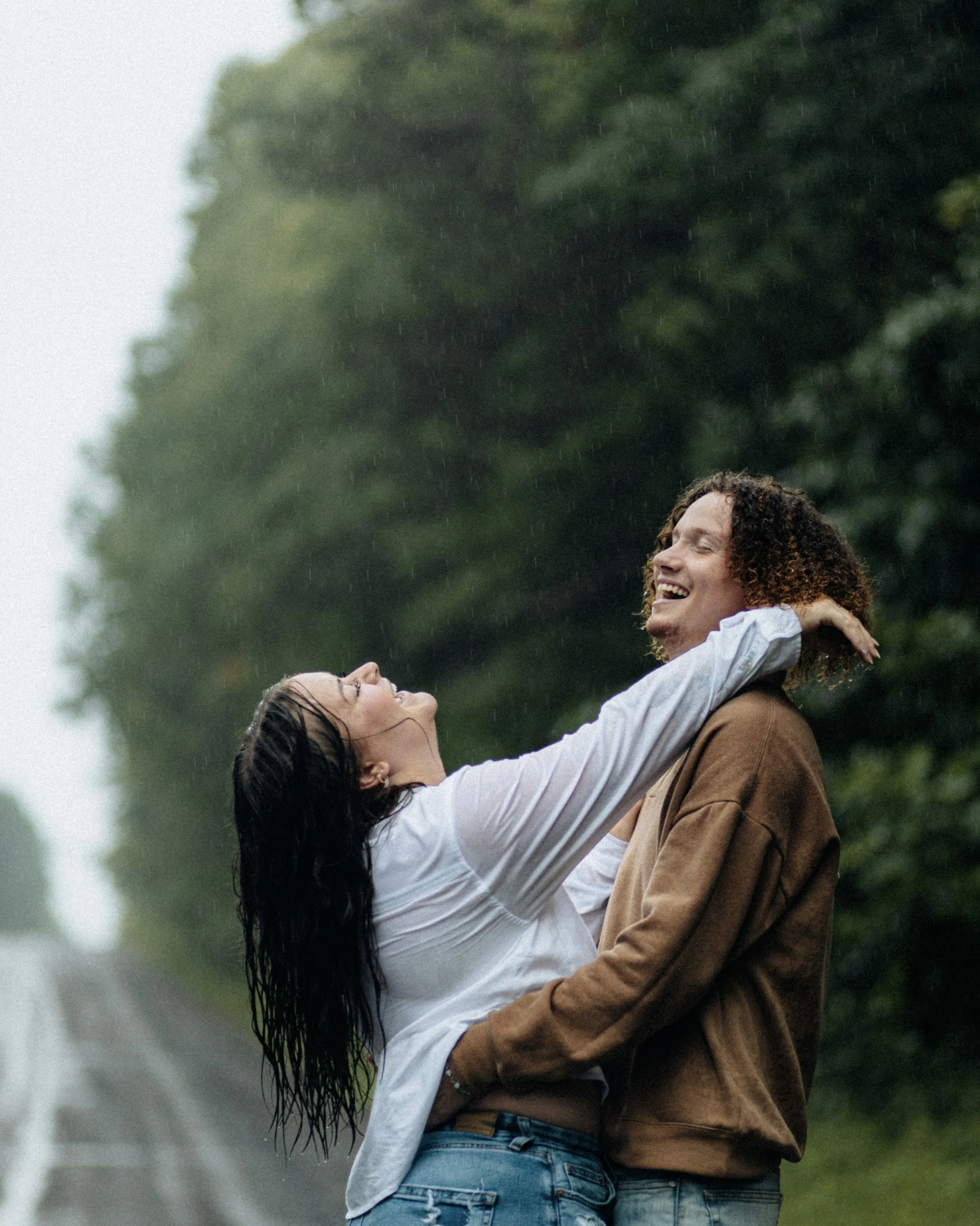 A couple happily embracing in the rain with lush green trees in the background.