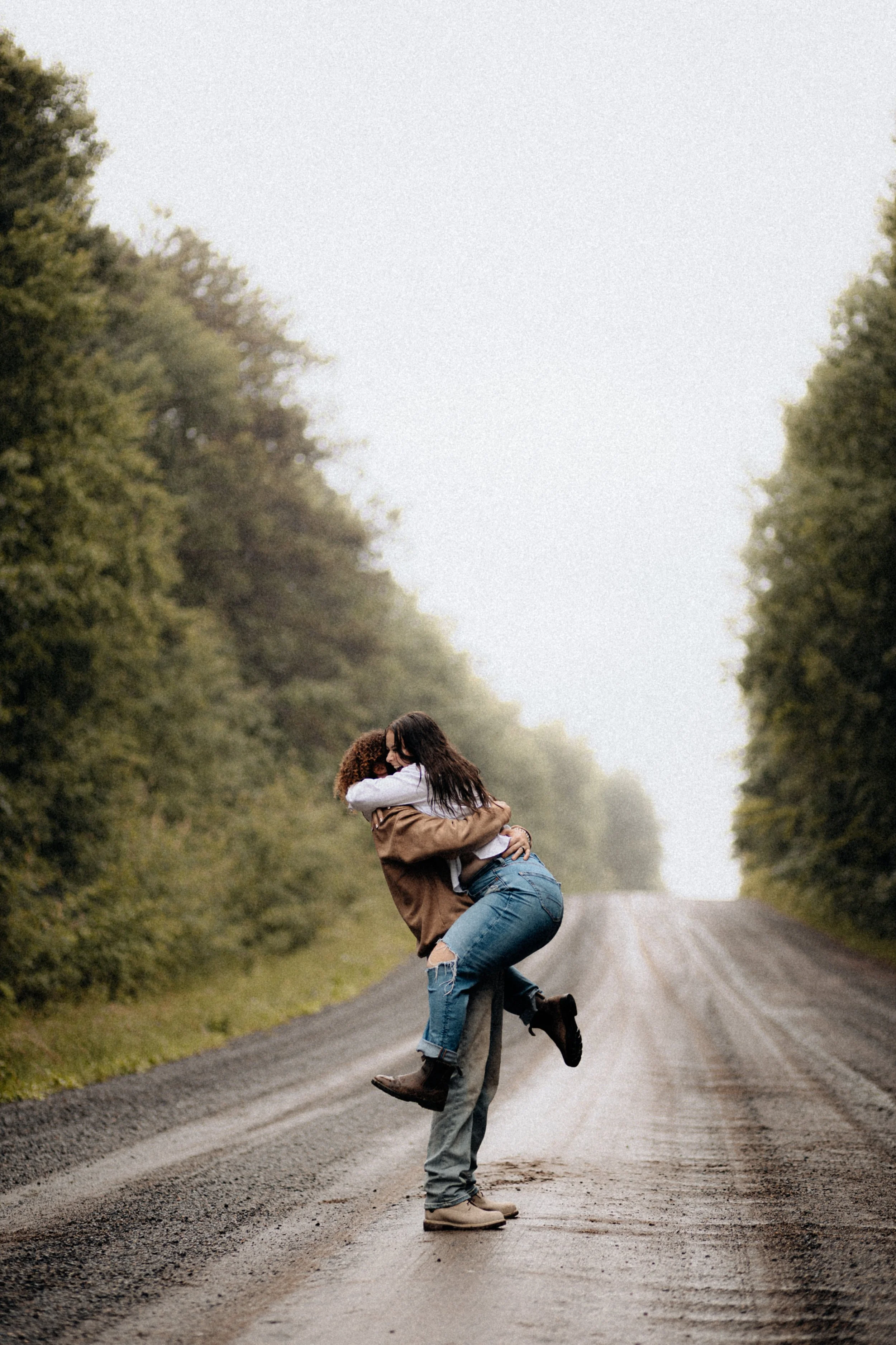 Two women hugging and laughing on a dirt road surrounded by trees.