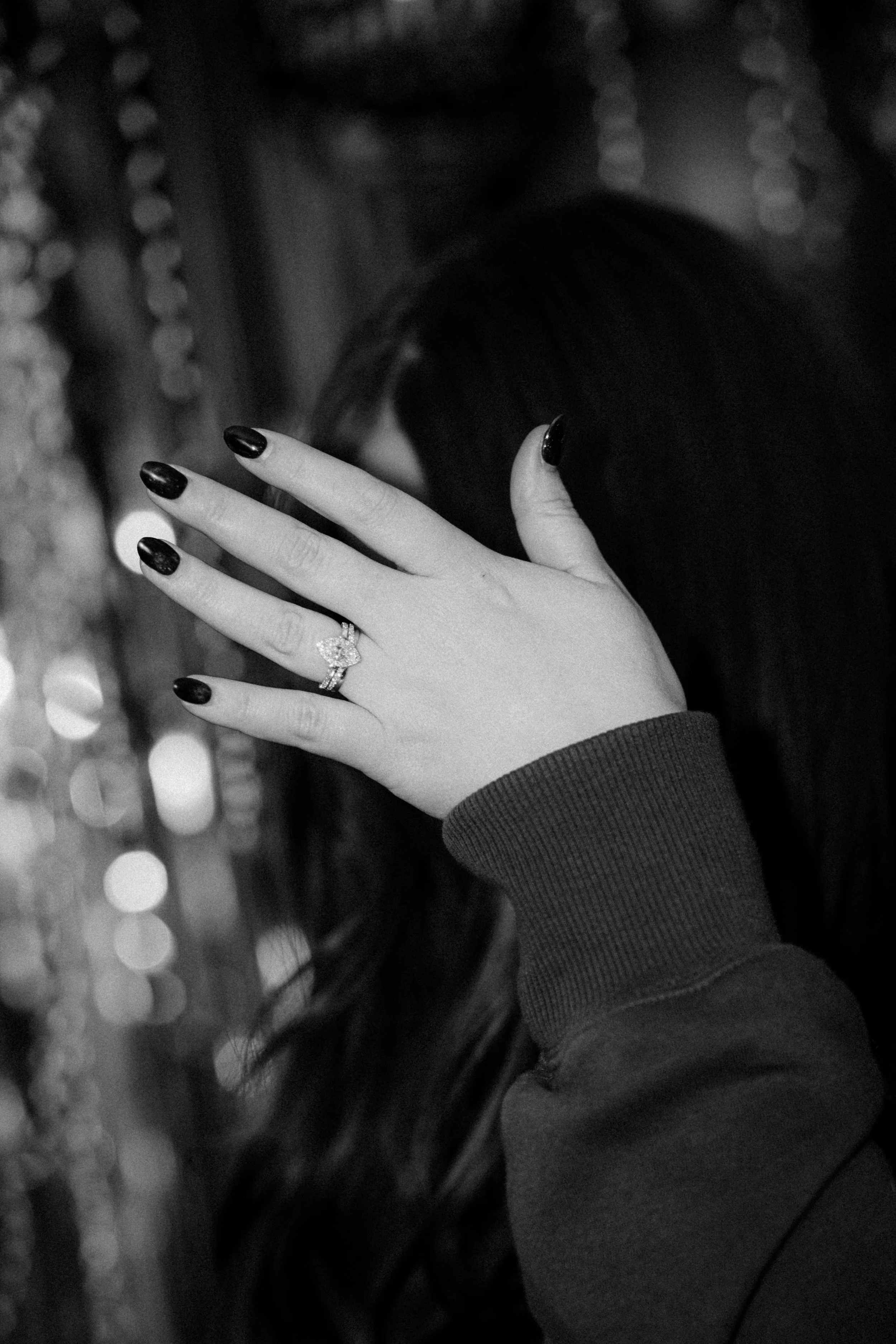 A woman covers her face with her hand, showing a ring on her finger, with dark painted nails, in a black-and-white photo.