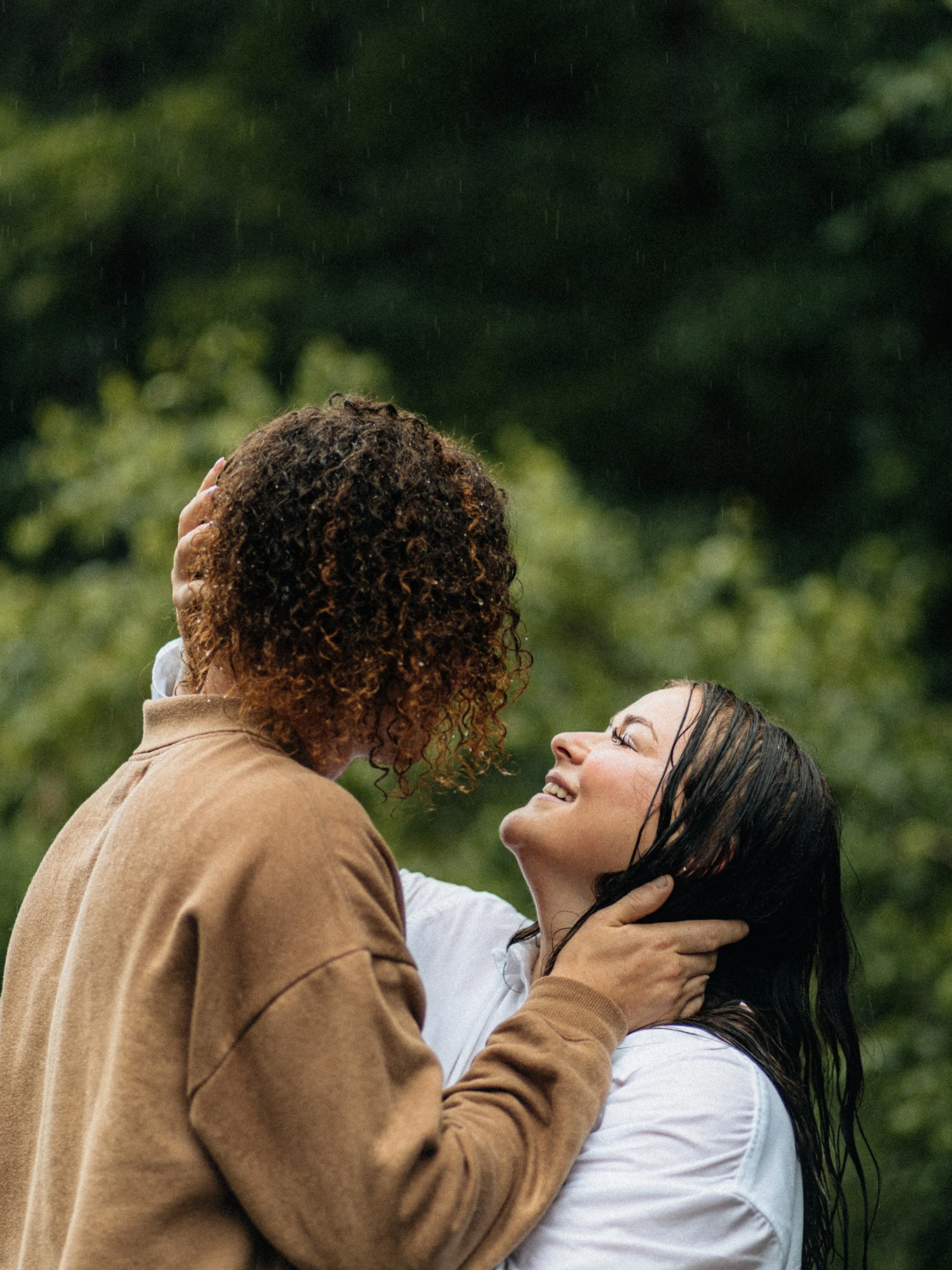 A tender moment between two women outdoors, with one woman smiling joyfully as she holds the other's face, rain falling, and a blurred green background.