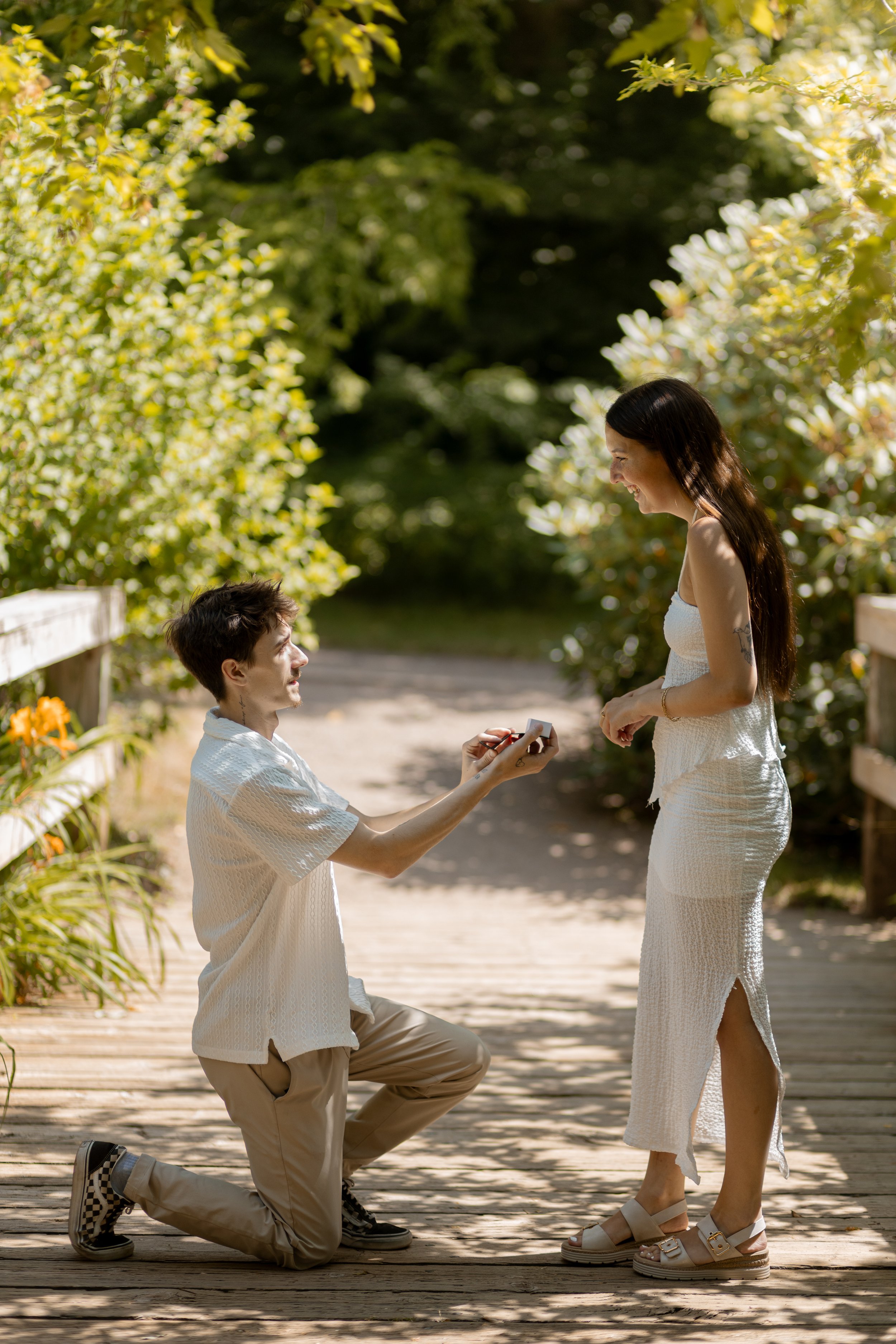 A man is kneeling on one knee on a wooden pathway, holding out a small box to a woman standing in front of him. The woman is smiling and looking at him, dressed in a white sleeveless dress. The scene is outdoors, surrounded by greenery and sunlight f