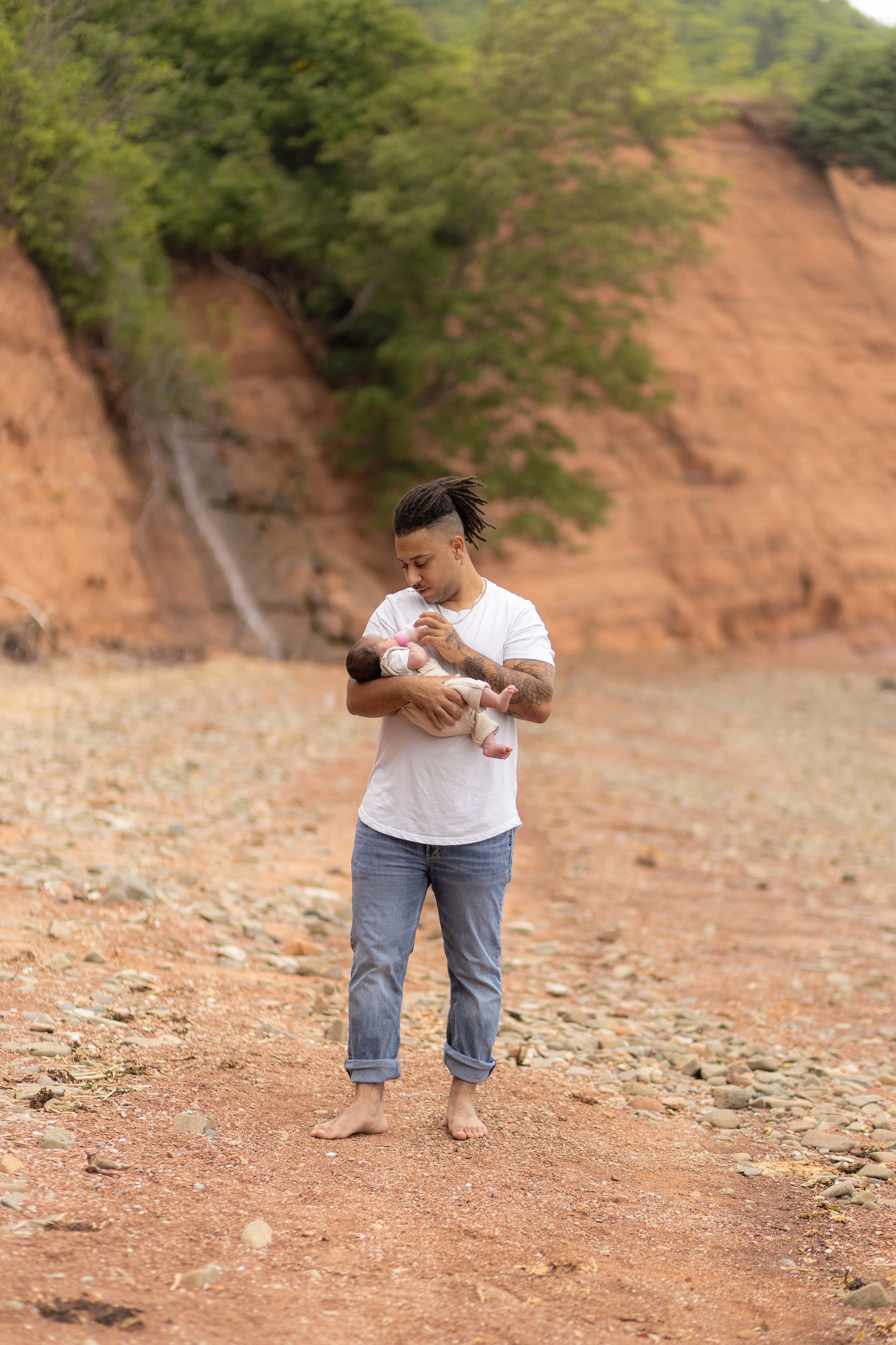 A man with dreadlocks, tattoos, and wearing a white t-shirt and rolled-up jeans, holding a baby in an outdoor natural setting with rocky terrain and trees in the background.