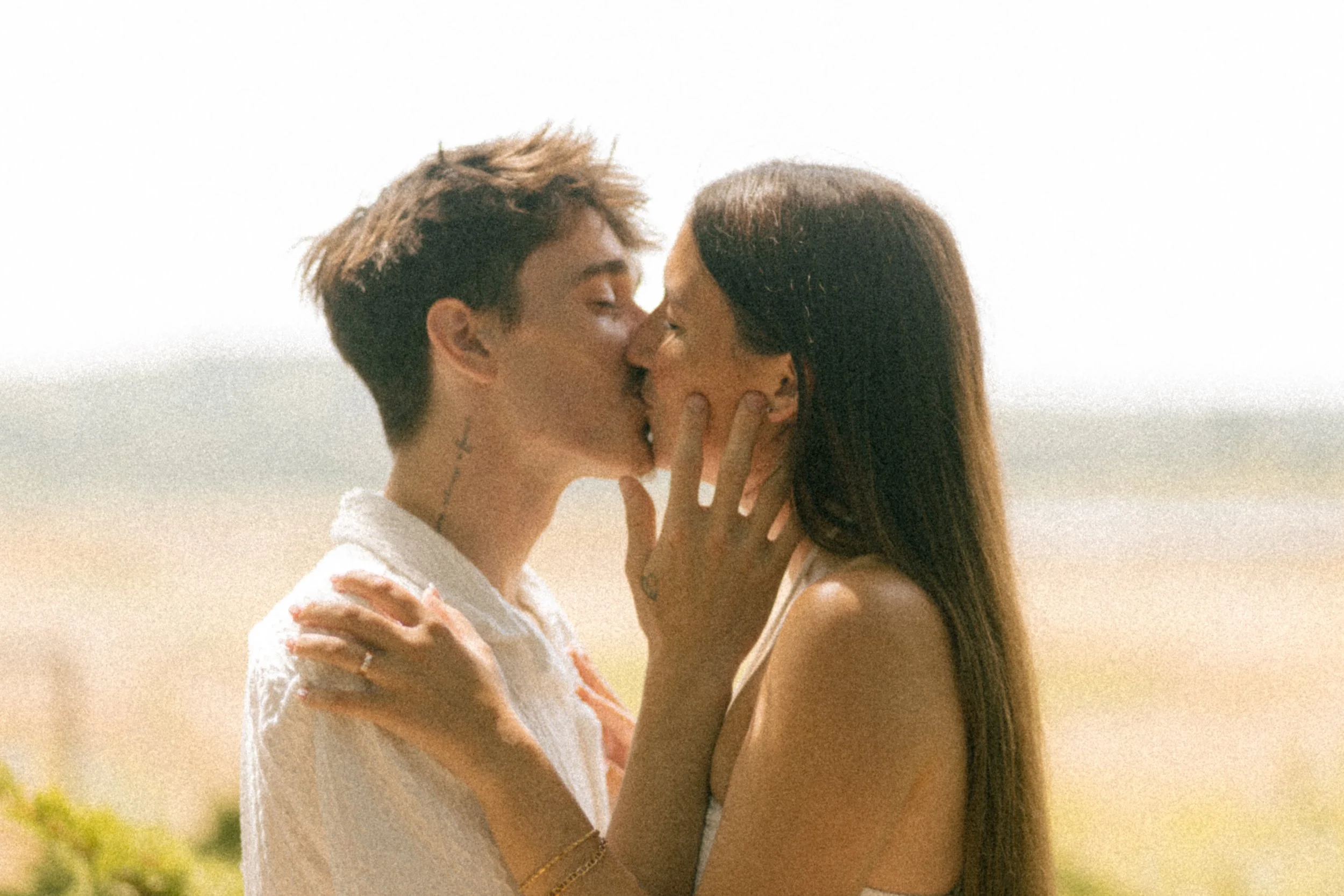A young couple sharing a kiss outdoors on a sunny day, with the ocean and sky in the background.