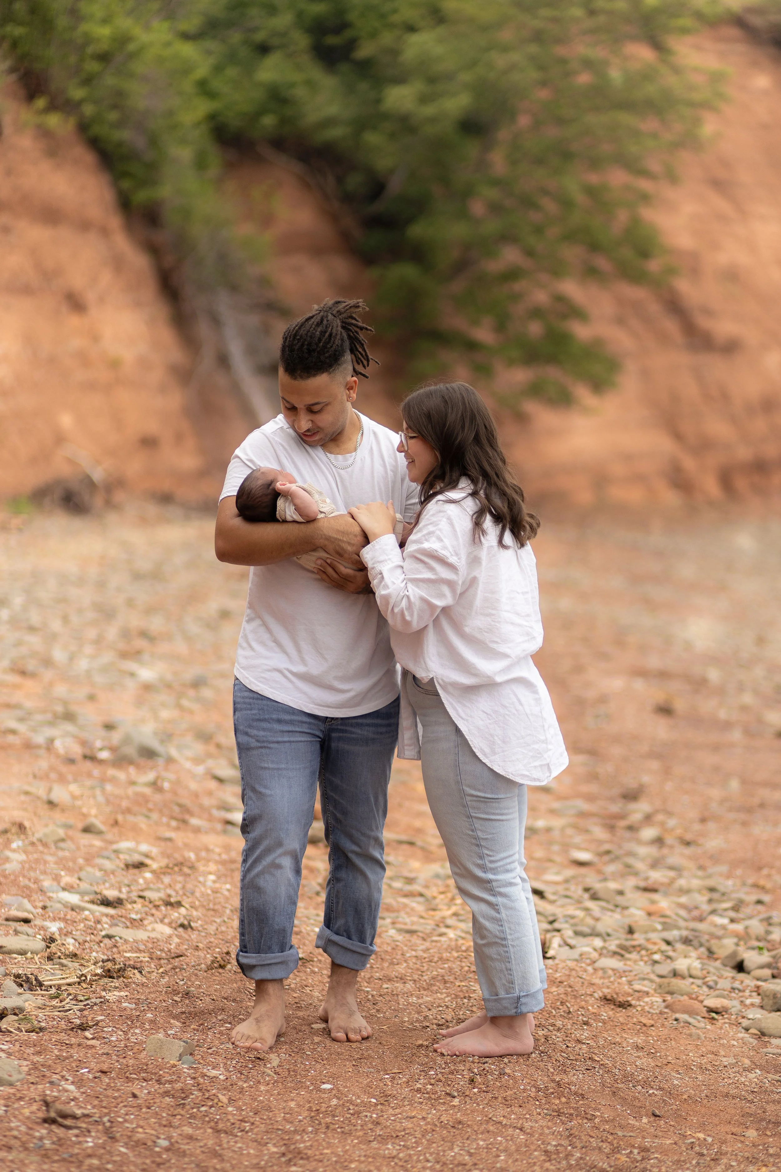 A family holding a newborn baby outdoors on a rocky, earthy landscape with a background of orange cliffs and green trees.