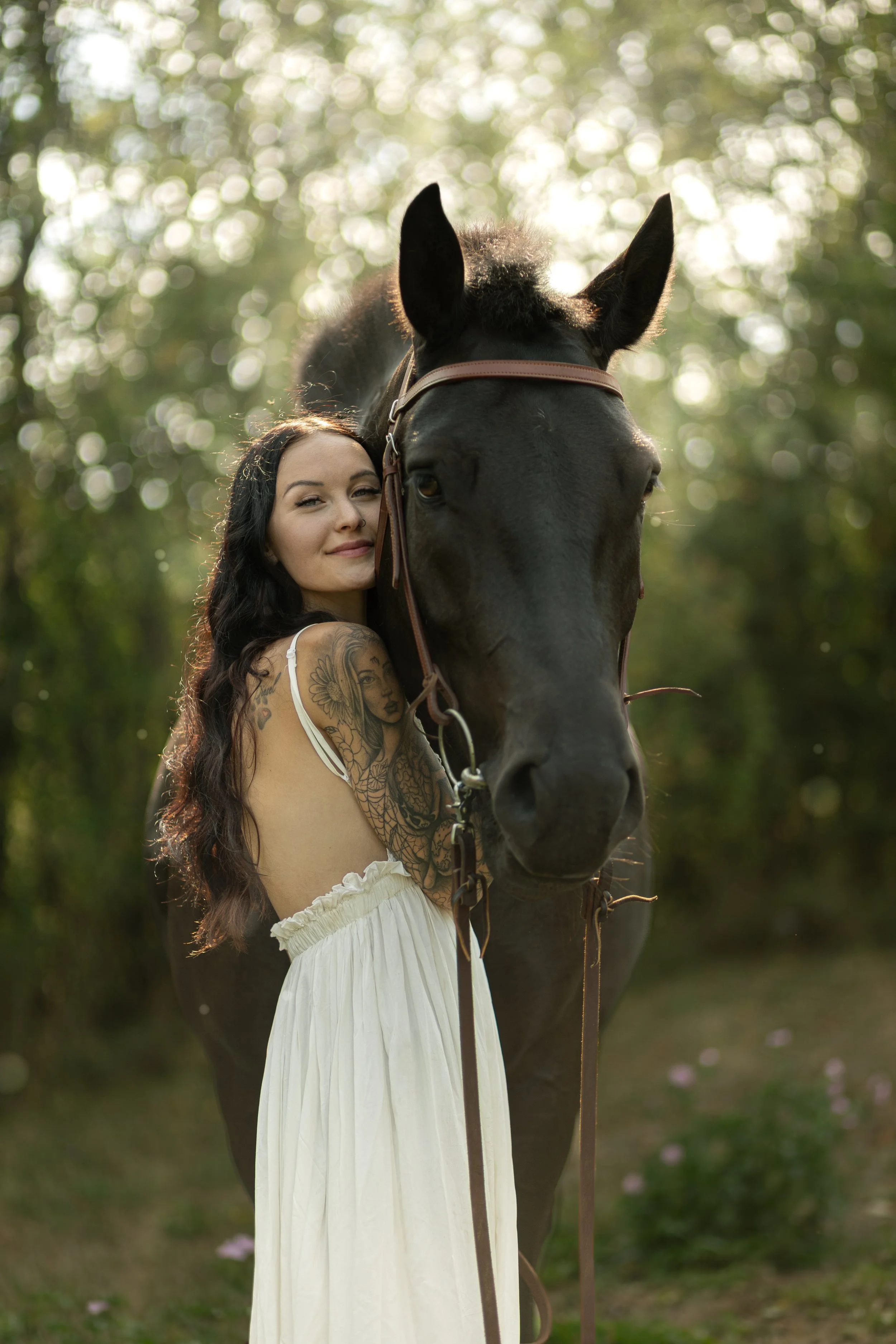 A woman with long dark hair and tattoos on her arm is standing closely beside a large black horse in a forested area, with sunlight filtering through the trees in the background.