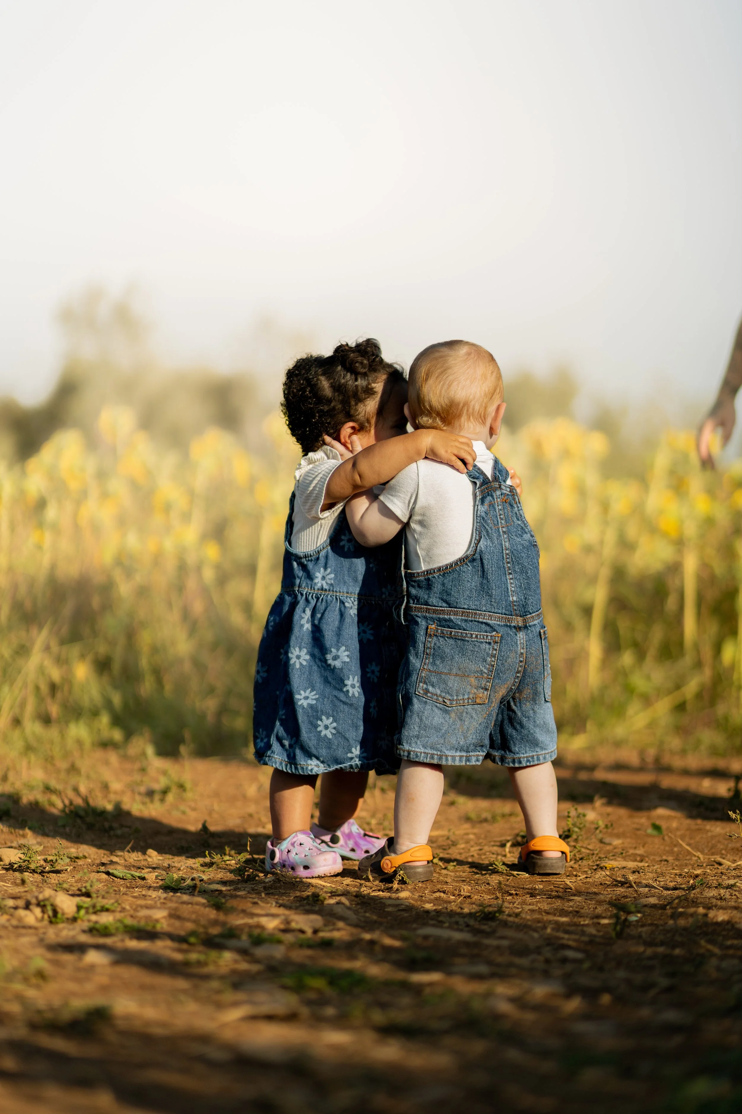 Two young children hugging outdoors on a dirt path with yellow flowers and green foliage in the background on a sunny day.