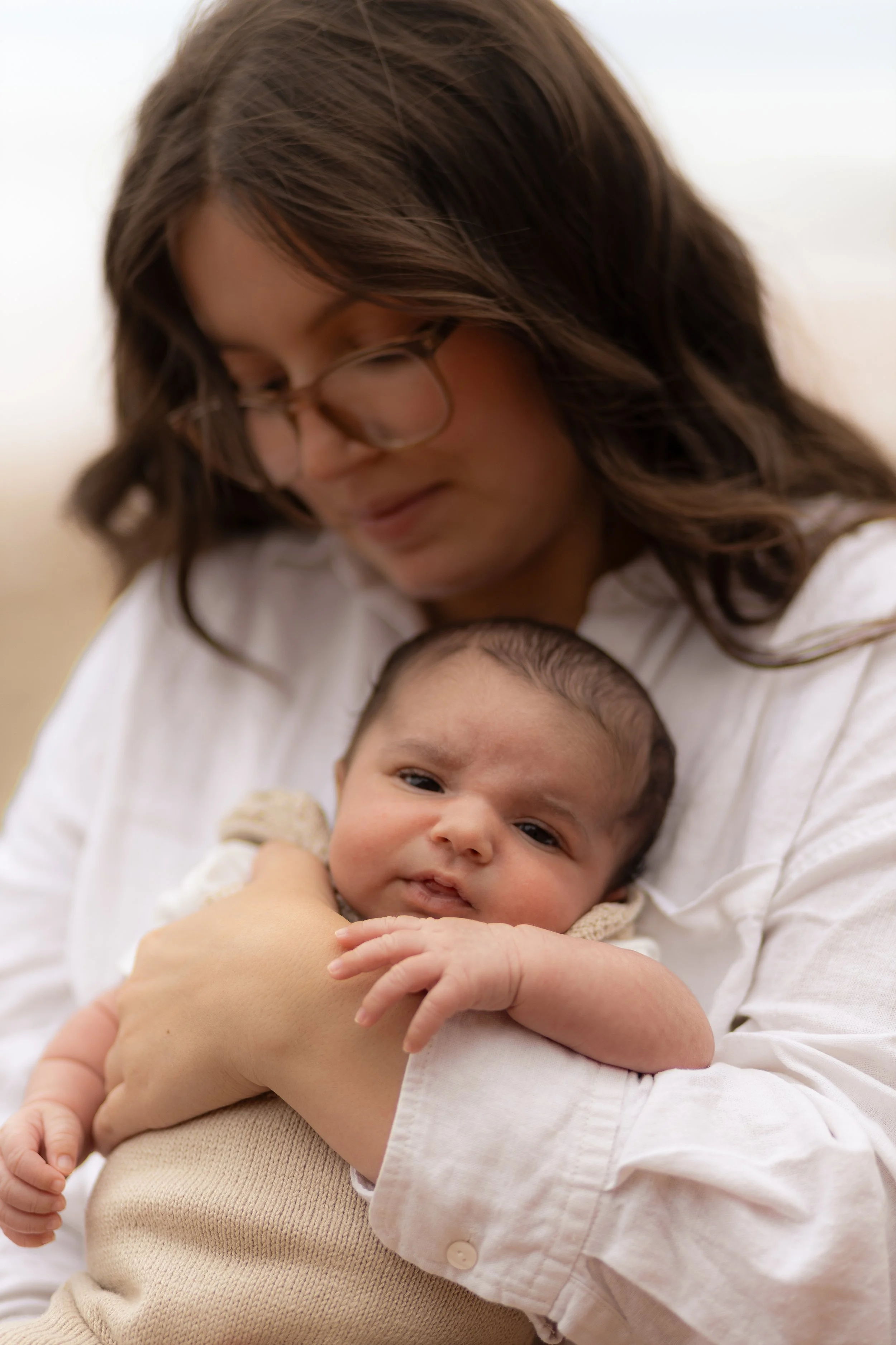 A woman holding a baby close to her chest, both wearing white tops, with the woman looking down at the baby in a soft, outdoor setting.