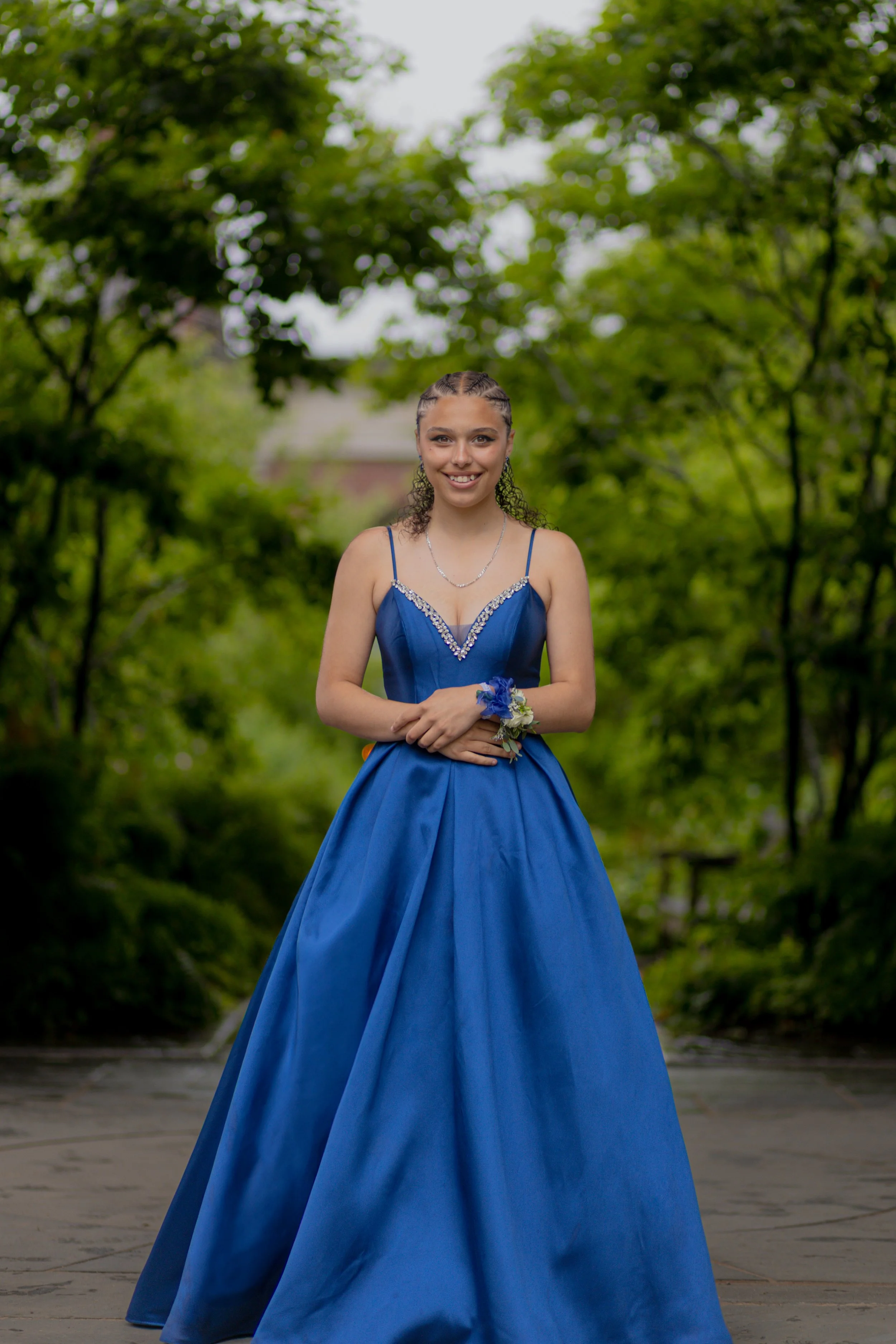A young woman in a blue formal dress standing outdoors with trees in the background.