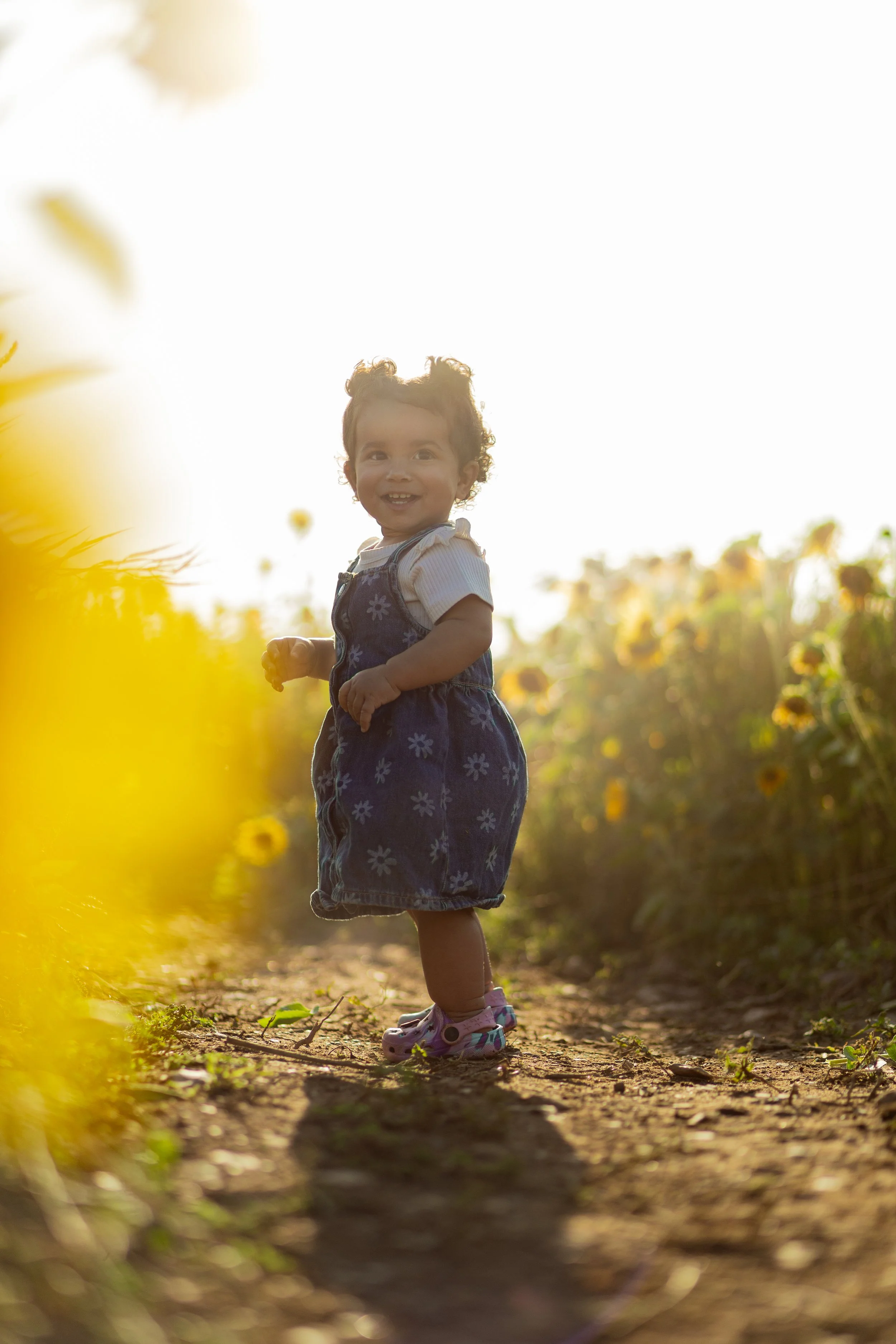 A young girl with curly hair smiling and walking on a dirt path in a sunflower field during sunset.