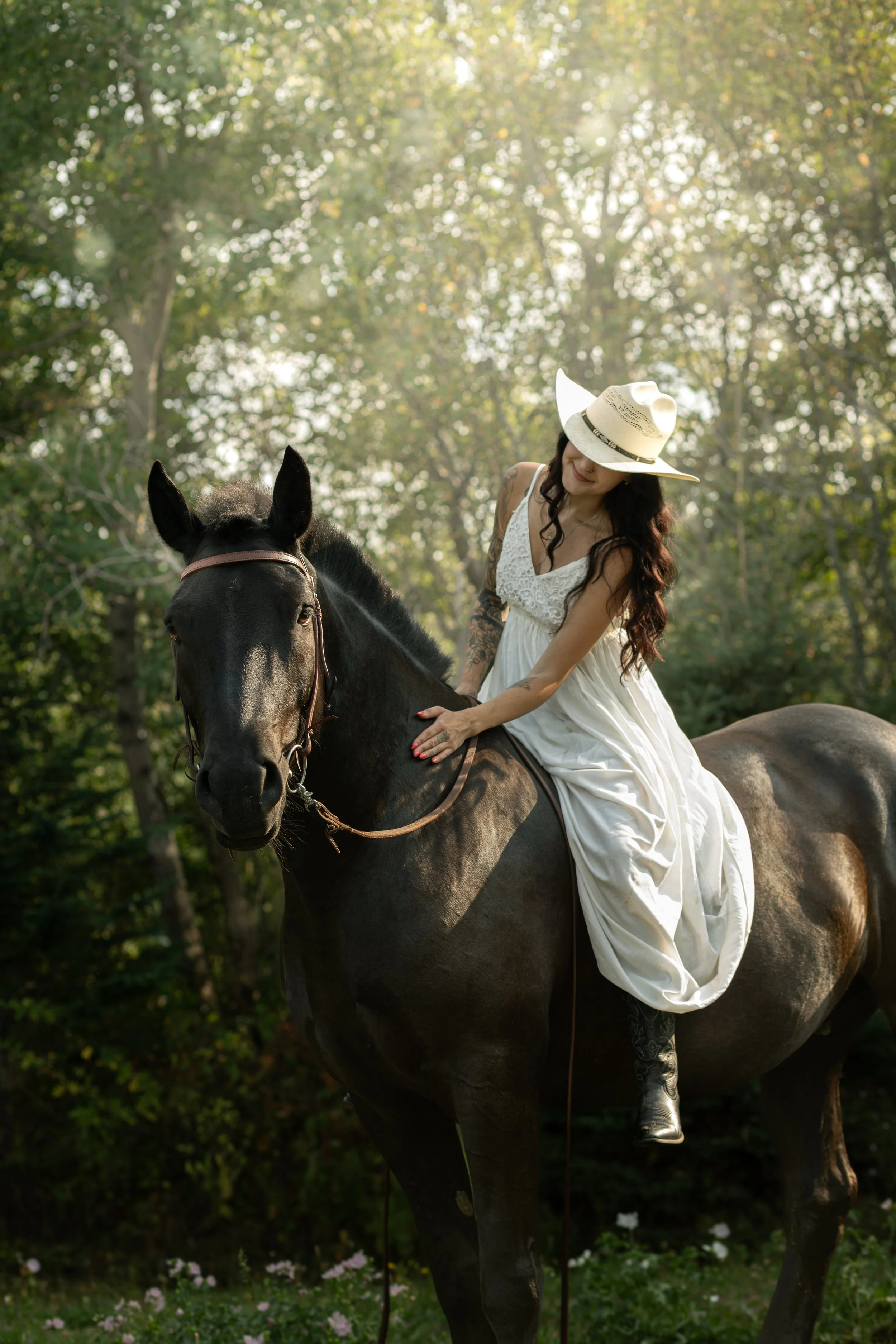 A woman wearing a white dress and wide-brimmed hat rides a black horse in a lush green forest.