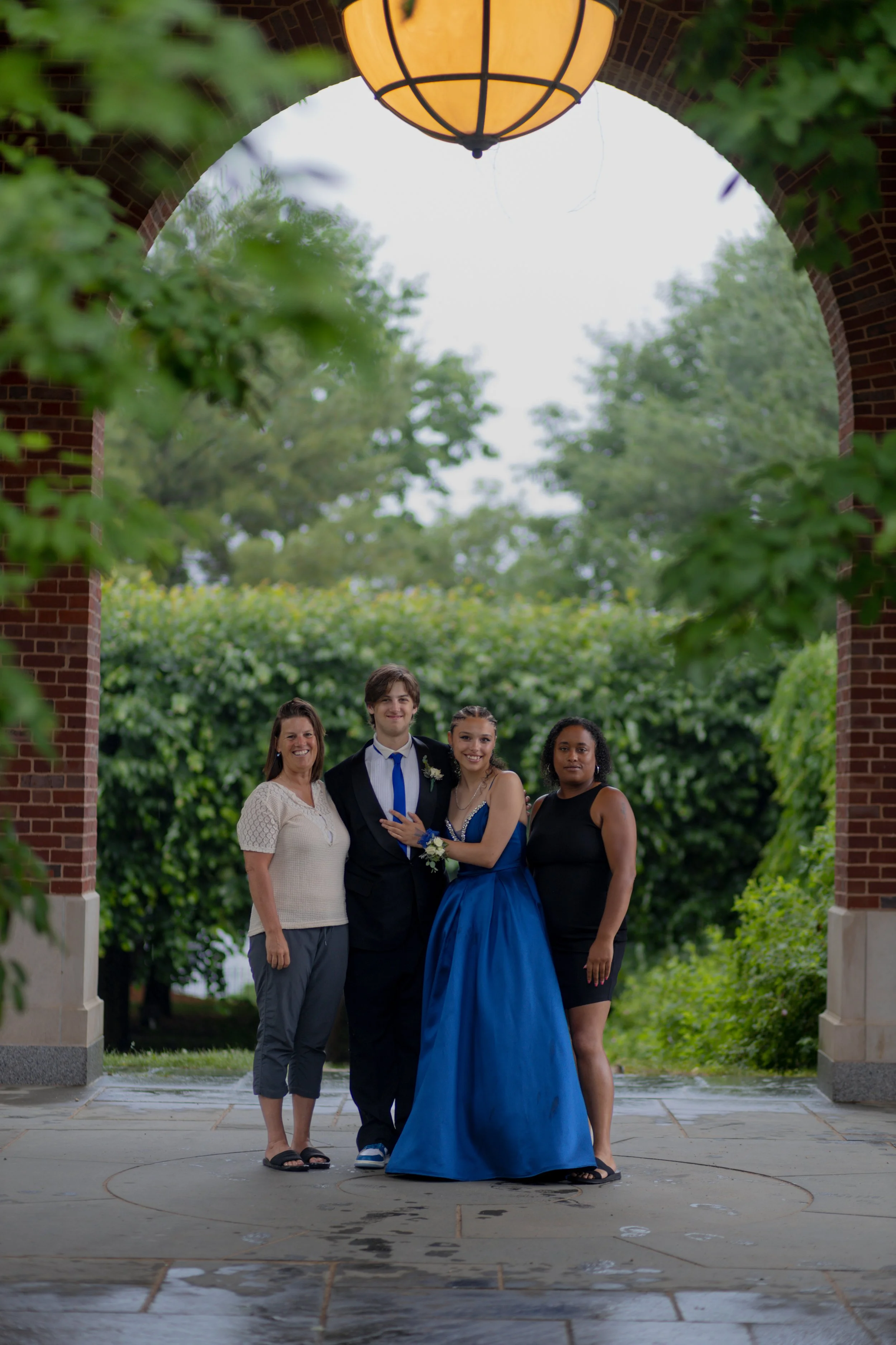 Four people standing under an archway, with the two in the middle dressed formally, possibly at a prom or wedding, and the woman on the right is wearing a long blue gown while the others are in casual attire. Green trees and bushes are in the backgro