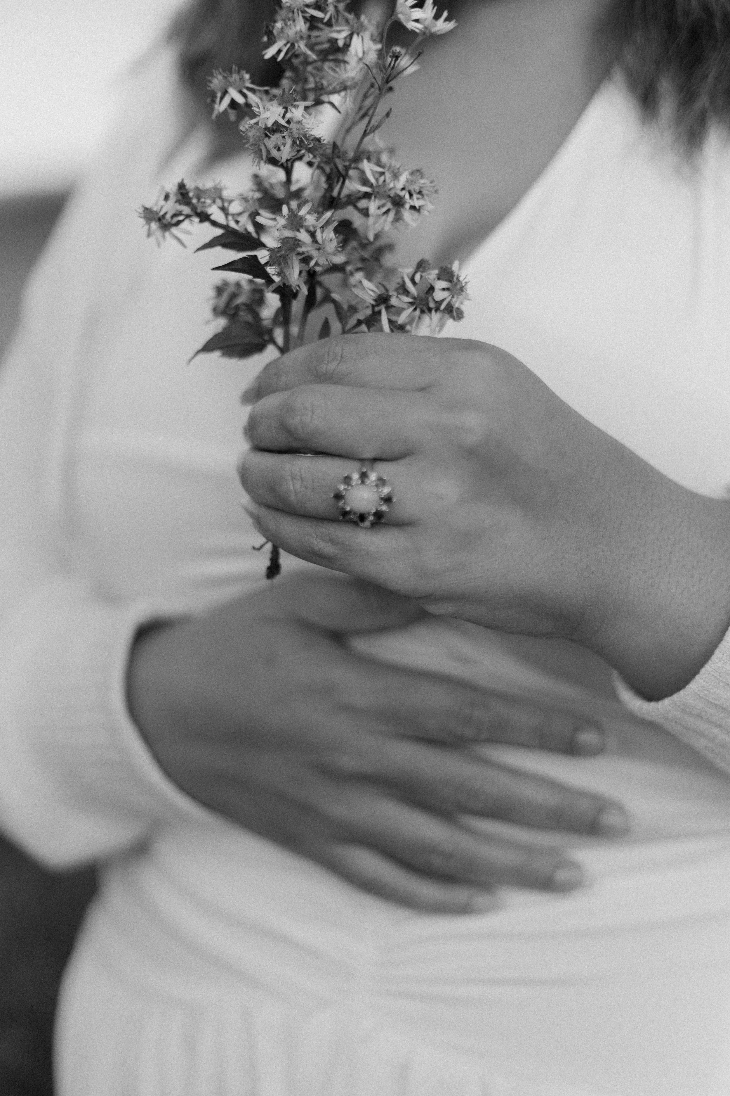 Close-up of a person holding a small bunch of wildflowers, wearing a large ring on their finger, with their hand placed over their chest. The image is black and white.