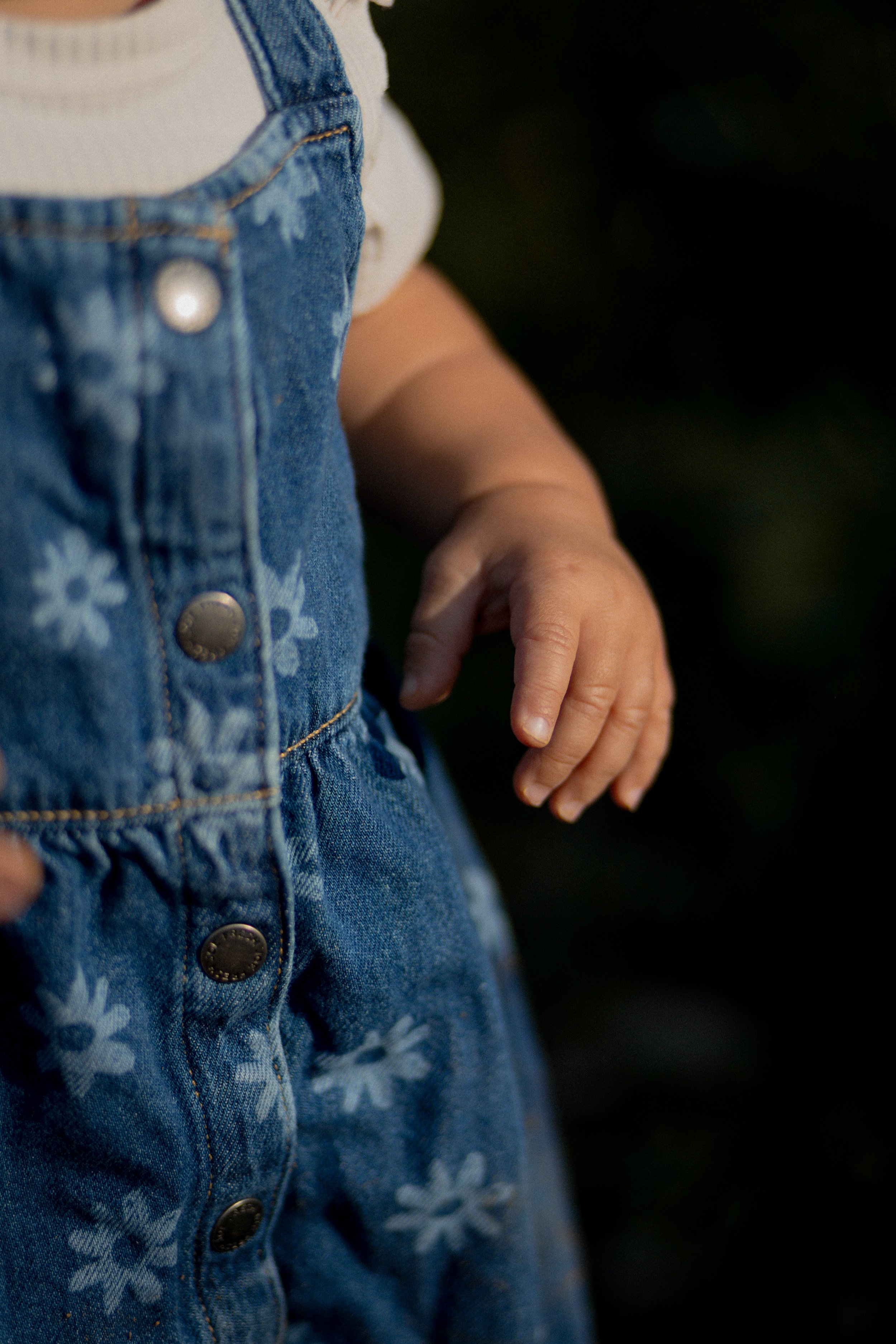 Close-up of a baby wearing denim overalls with a floral pattern and a white shirt, with one hand visible.