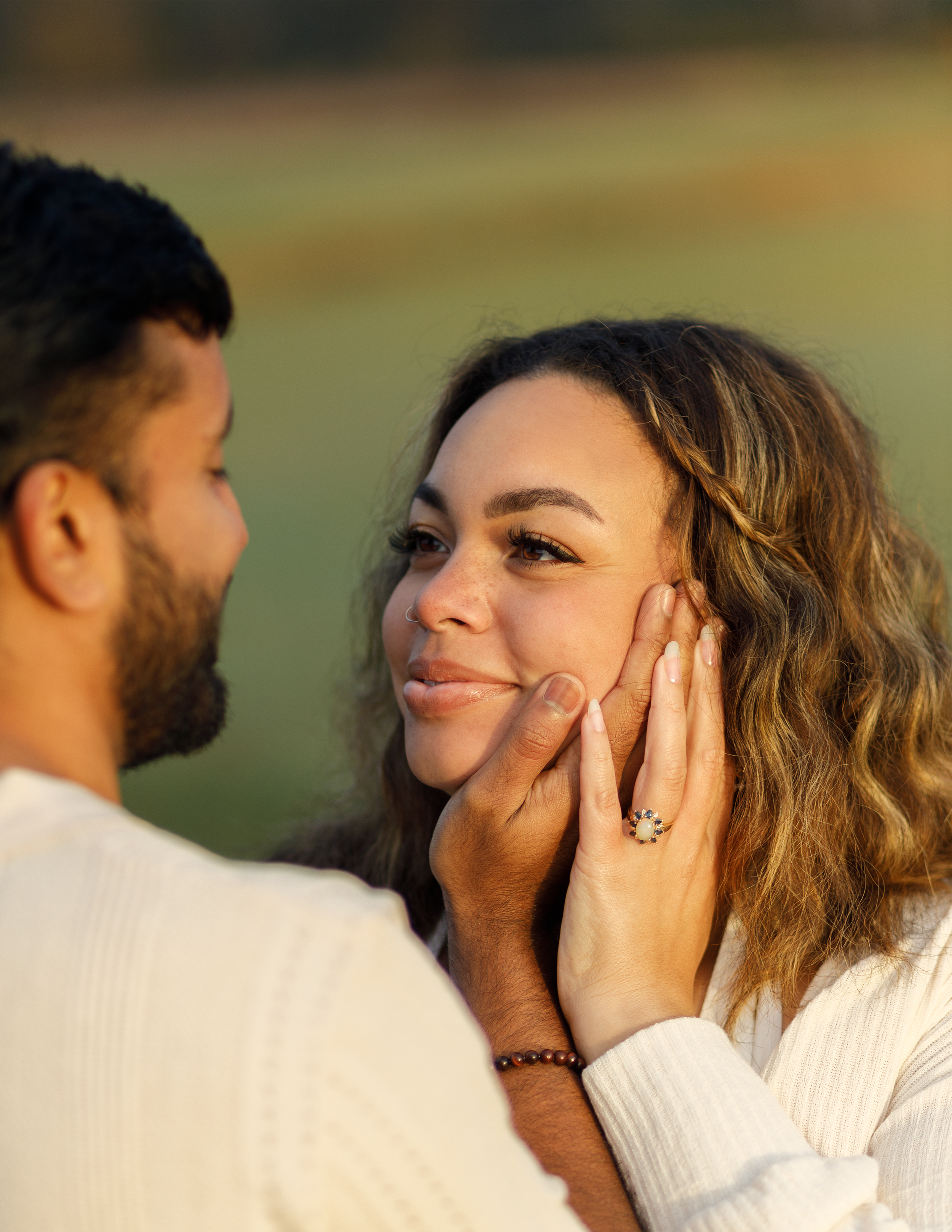 A couple sharing an intimate moment outdoors, with the man gently holding the woman's face and looking into her eyes, both smiling softly.