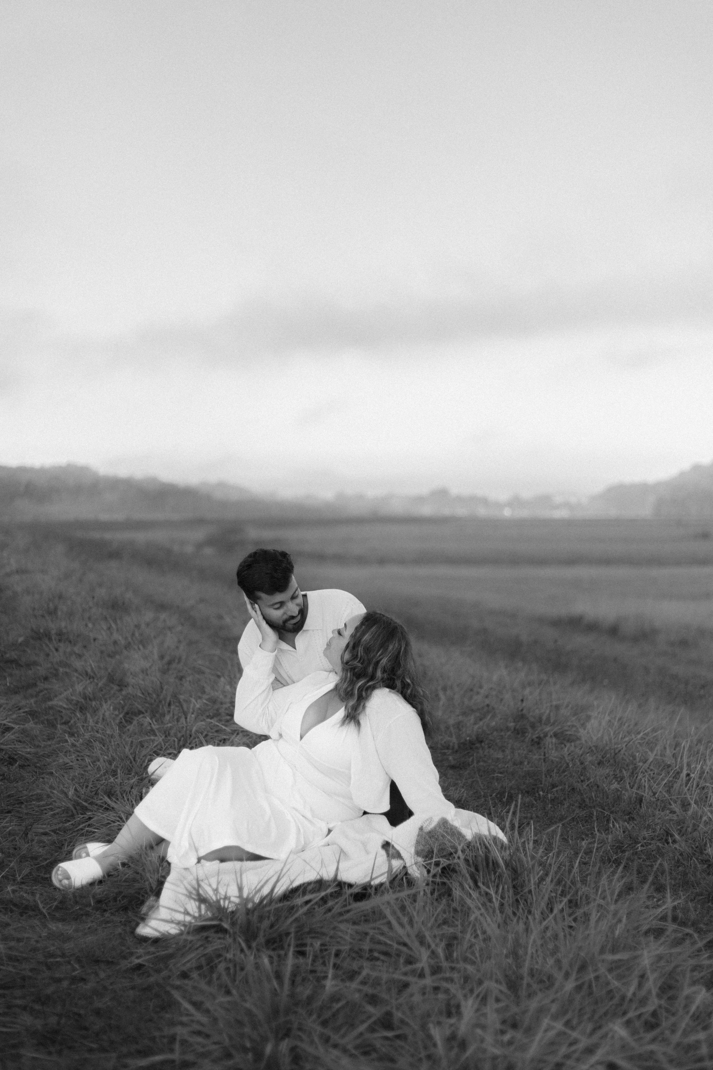 A black-and-white photo of a couple sitting in a grassy field, embracing and looking into each other's eyes, with rolling hills and an overcast sky in the background.