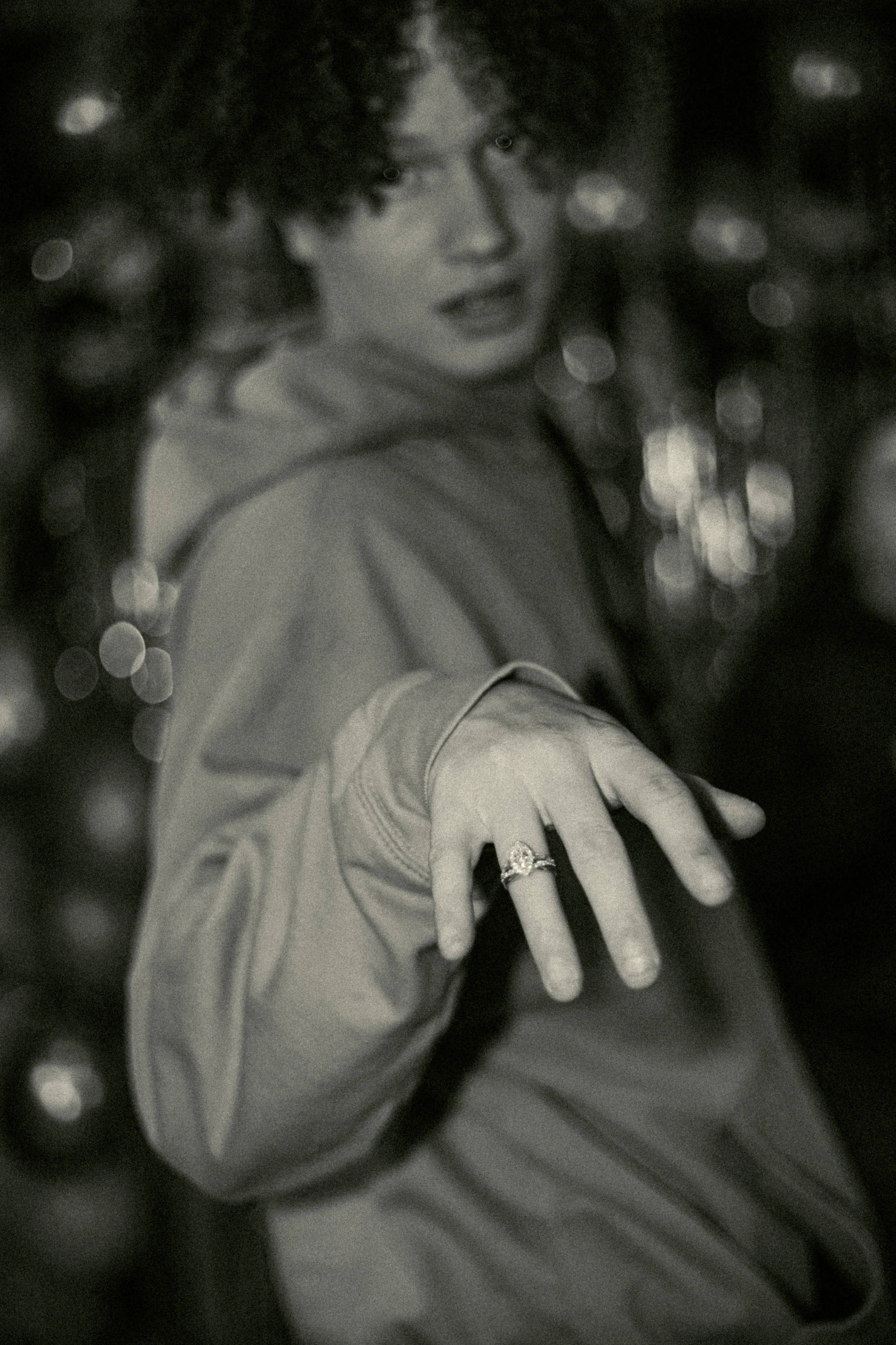 A person with curly hair is extending their hand towards the camera, wearing a ring, with a blurred background of lights.