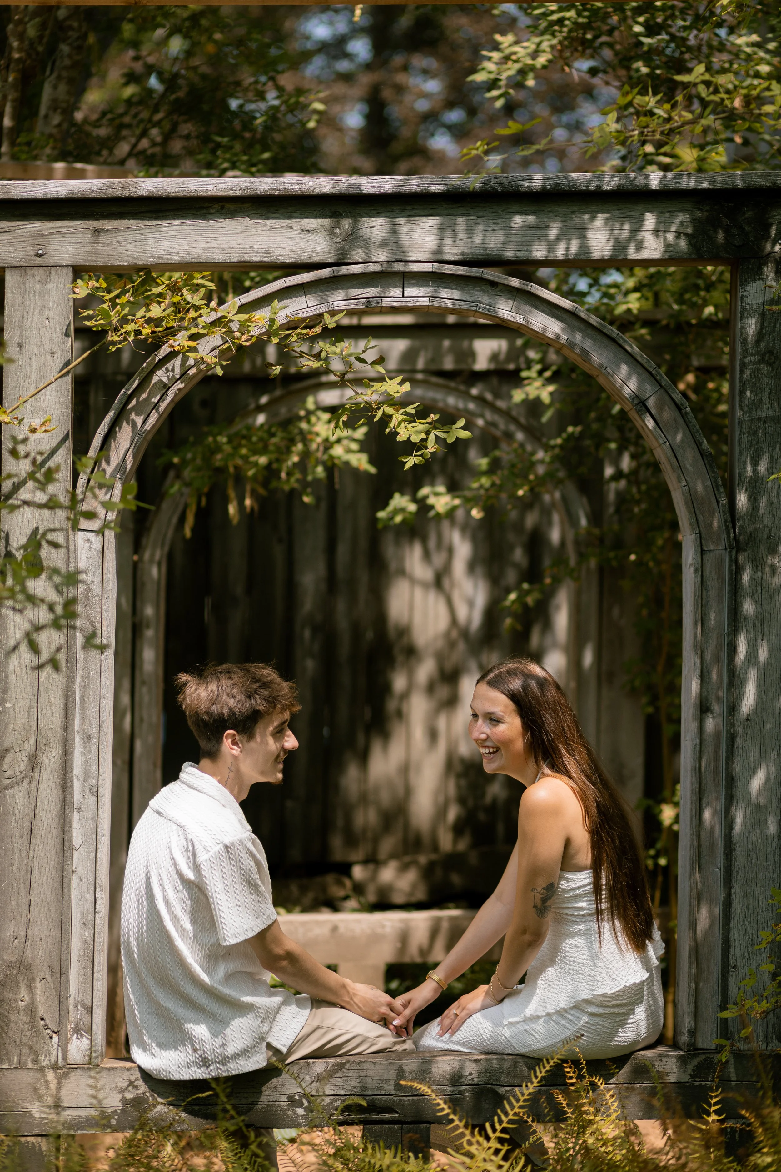 Two women sitting on a wooden window frame outdoors, smiling and holding hands, surrounded by greenery.