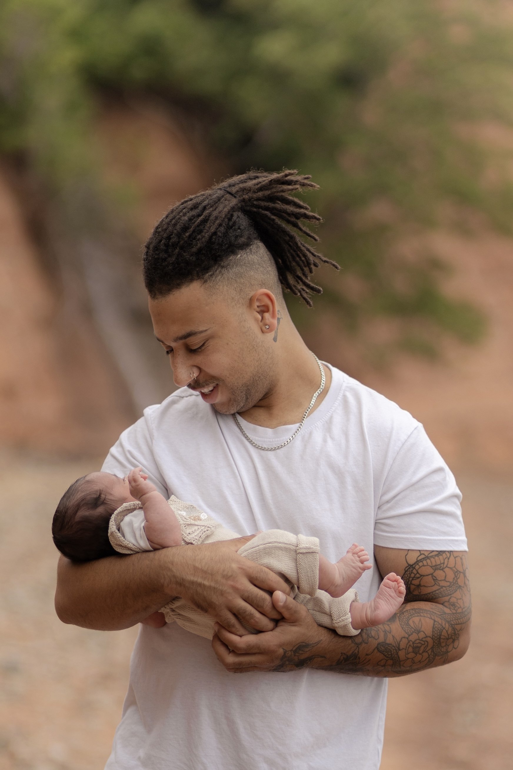 A man with tattoos and dreadlocks holding a newborn baby outdoors, smiling and looking down at the baby.