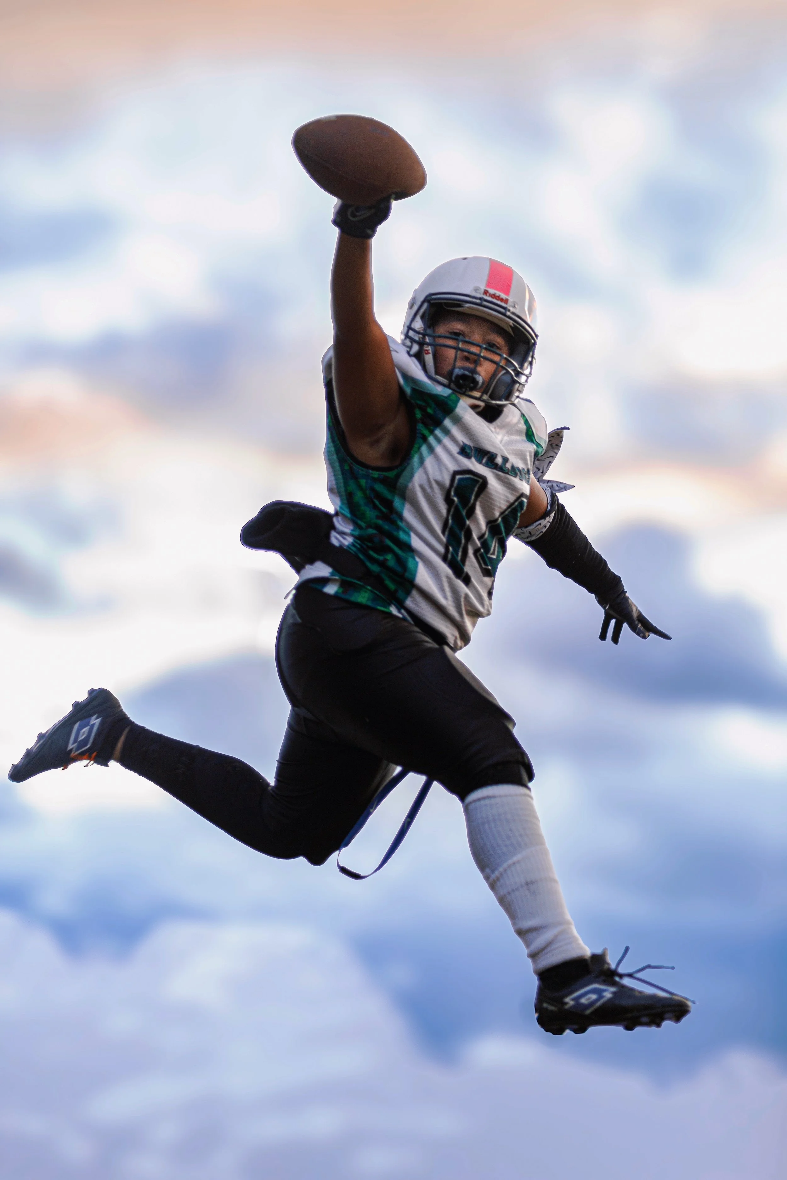 A football player in a gray and teal uniform is leaping in the air, holding a football in his right hand, with a cloudy sky in the background.