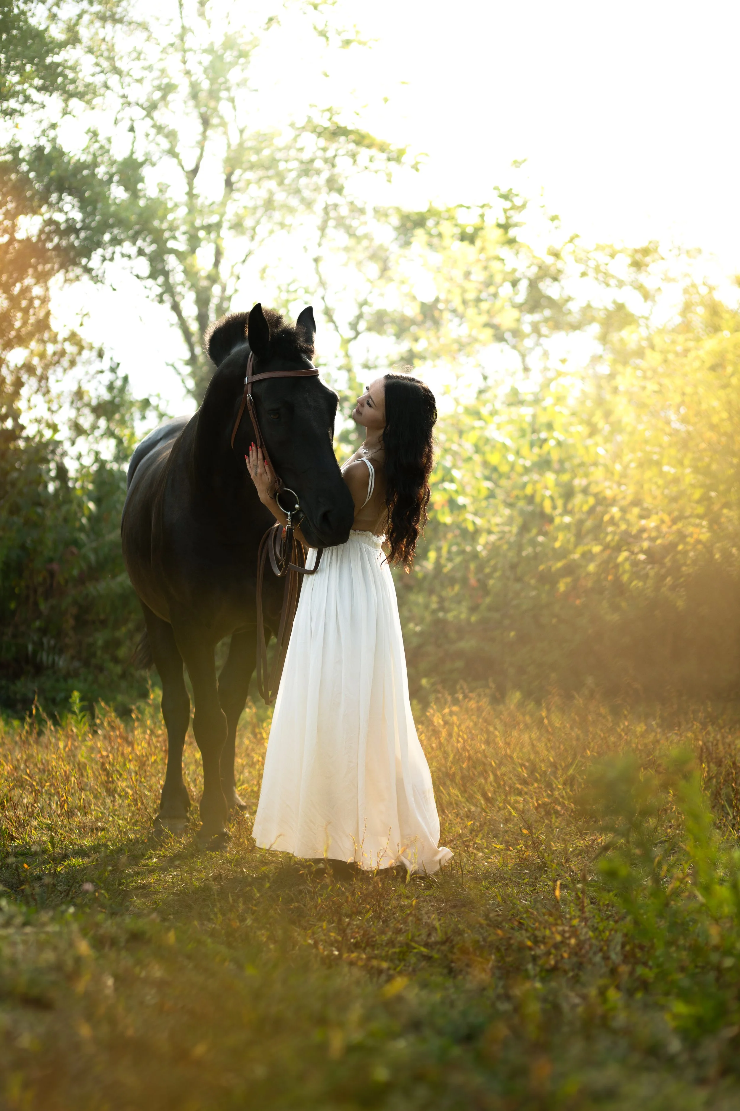Woman in a white dress standing next to a black horse in a sunlit outdoor setting with trees and golden light.