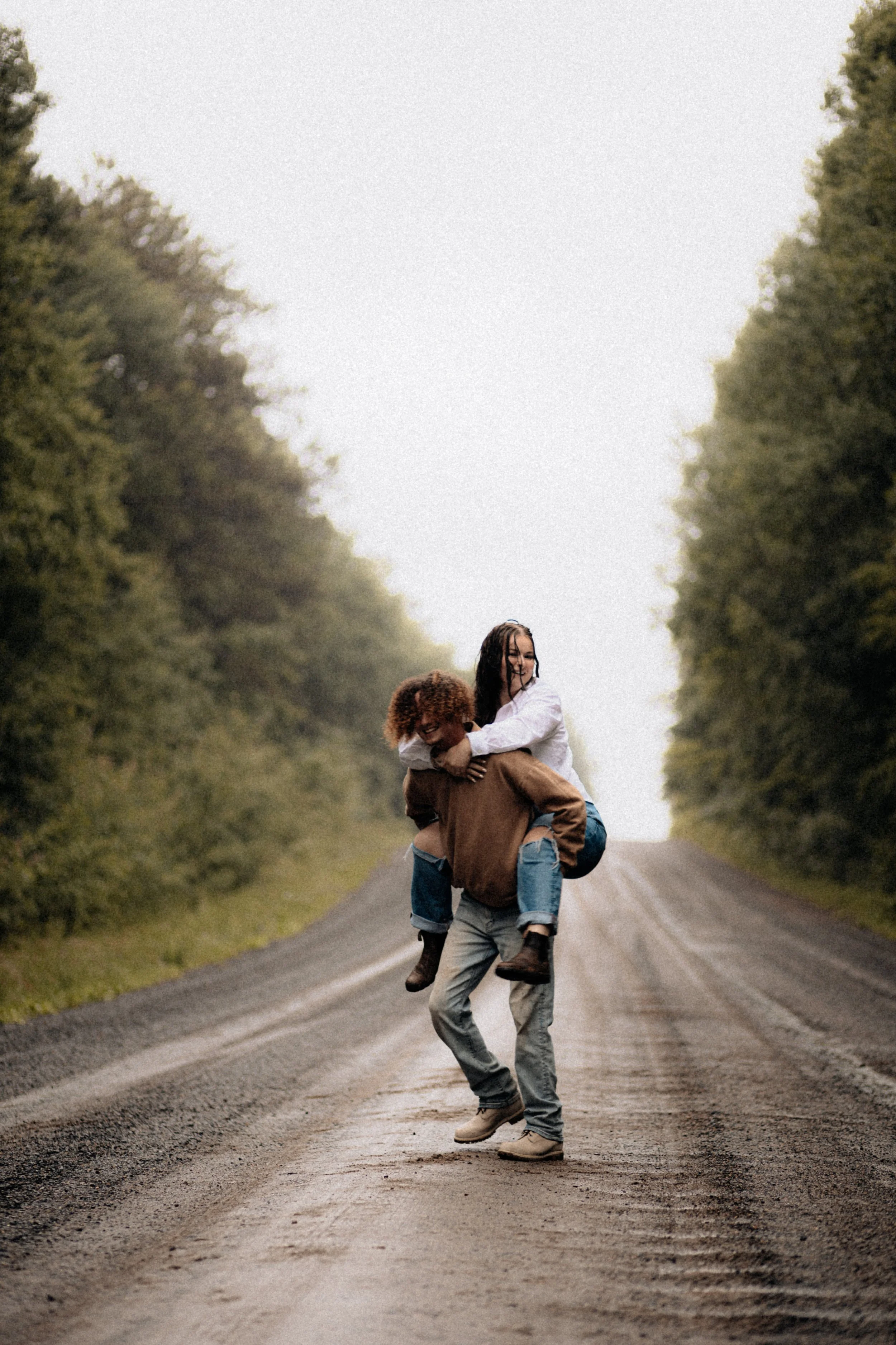 Three people, two women and one man, enjoying a piggyback ride on a dirt road surrounded by trees, with an overcast sky.