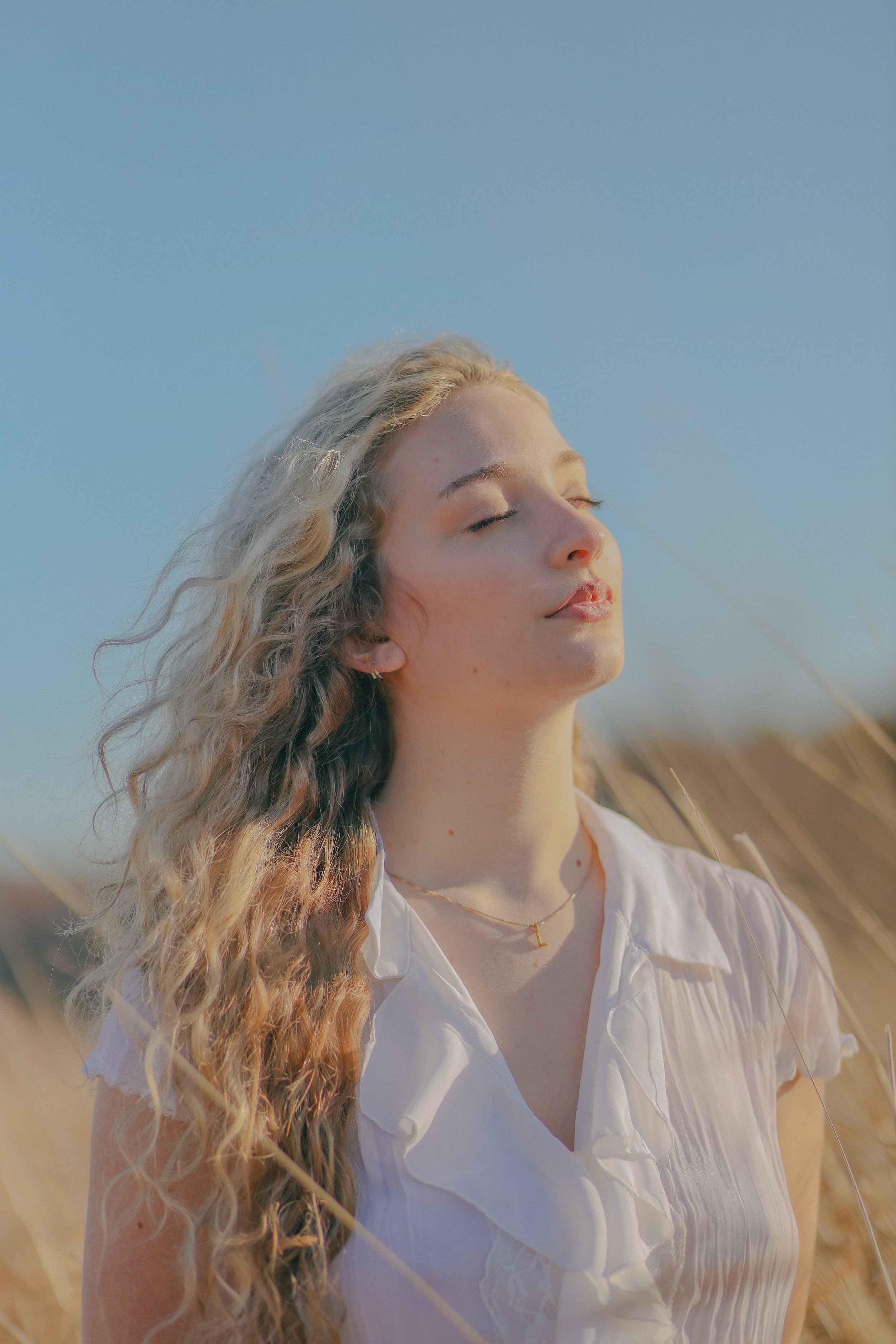 A young woman with long, wavy blonde hair and fair skin, wearing a white blouse and a delicate gold necklace, standing with her eyes closed in a sunny field of tall, golden grass with a clear blue sky in the background.