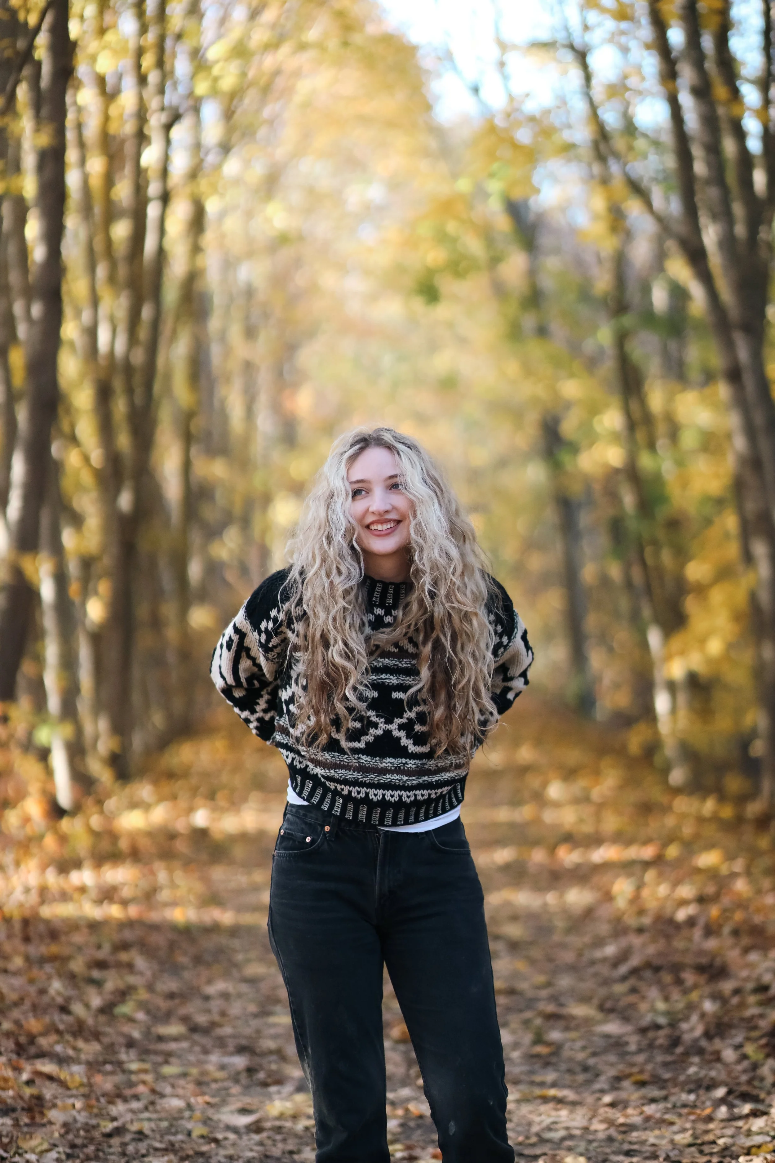 A young woman with long curly blonde hair smiling and standing on a leaf-covered path in a forest with autumn foliage.