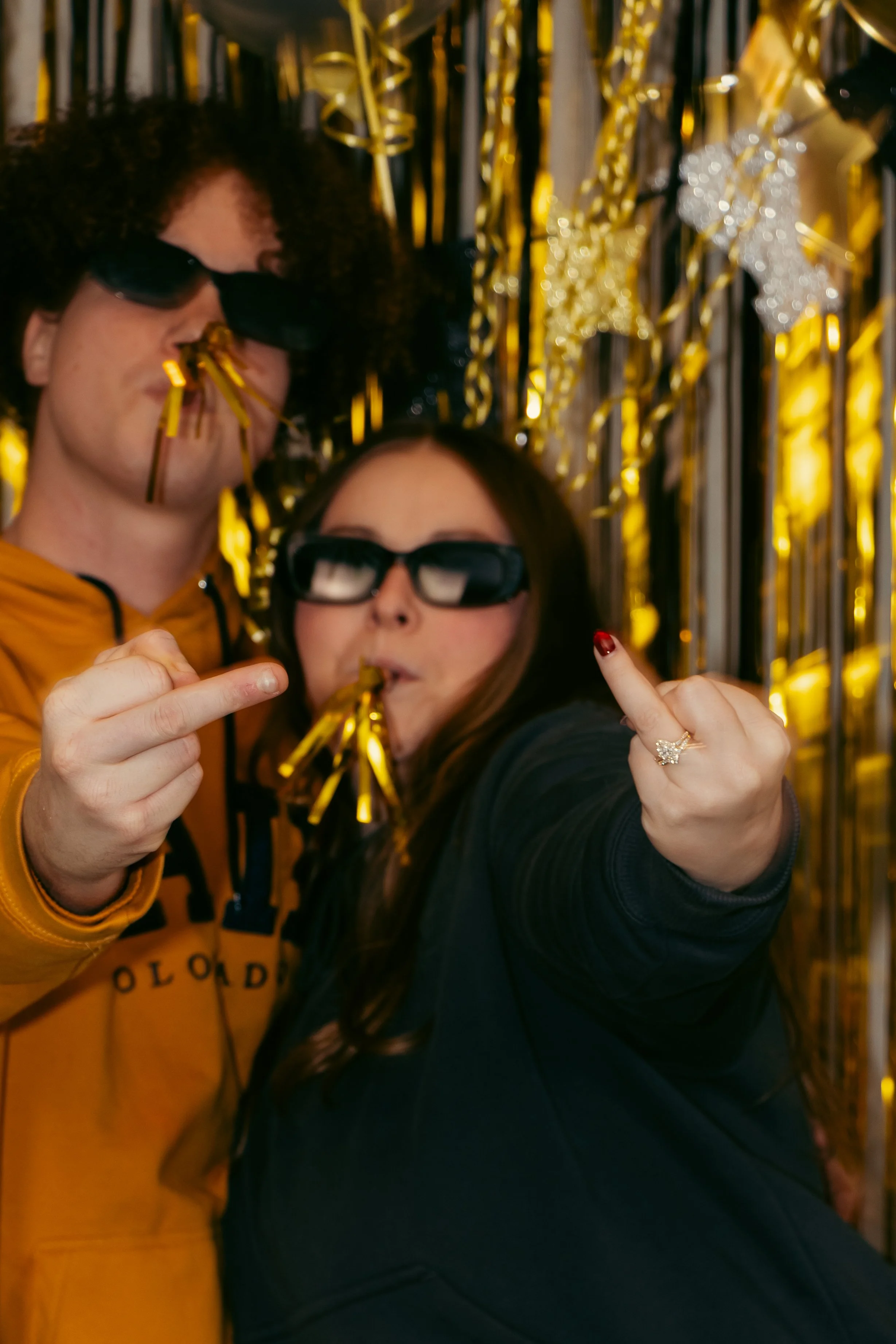 Two people celebrating New Year's Eve with party glasses and golden confetti in front of a golden decorations background.