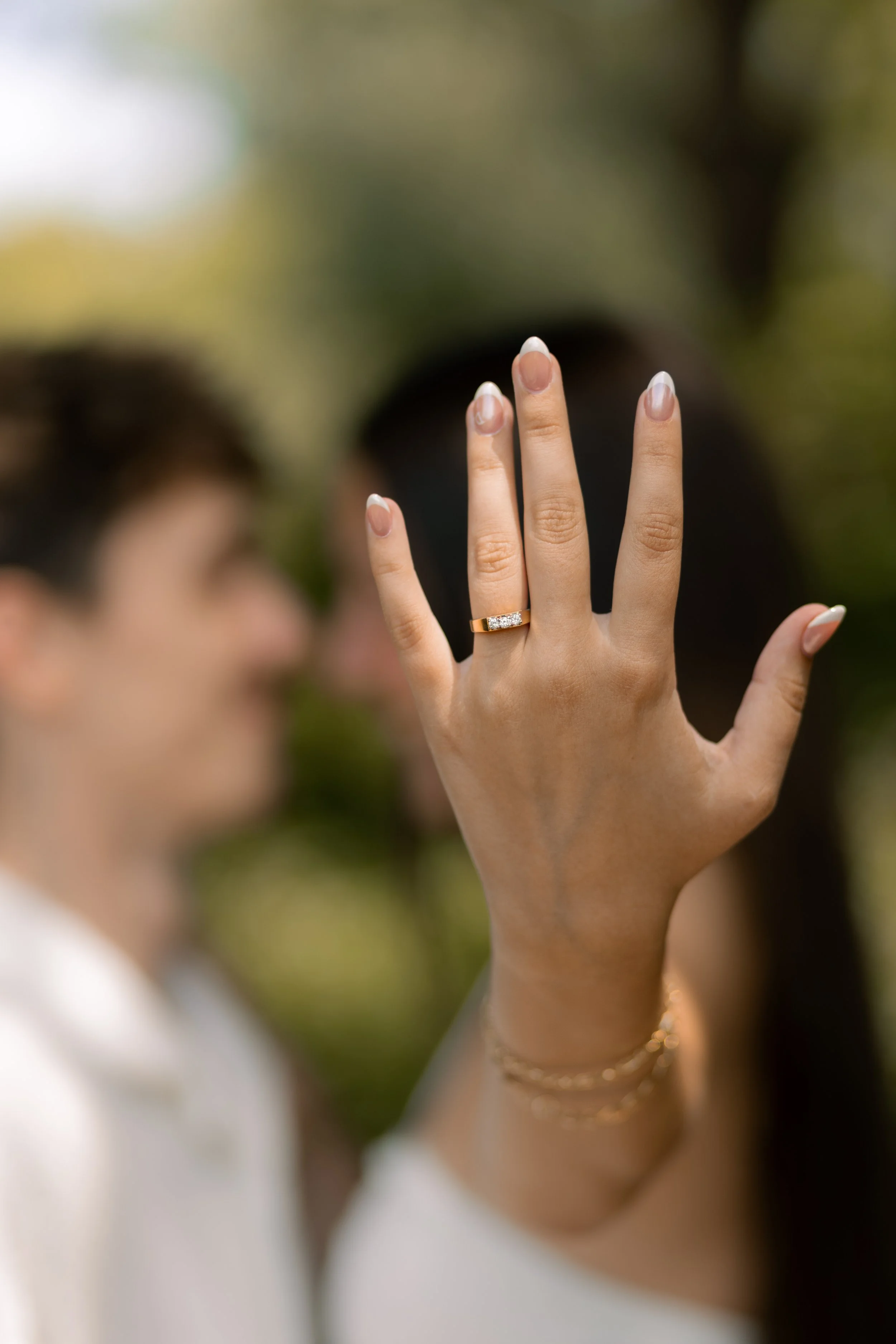 Close-up of a woman's hand showing an engagement ring with a diamond band, with blurred couple in the background.