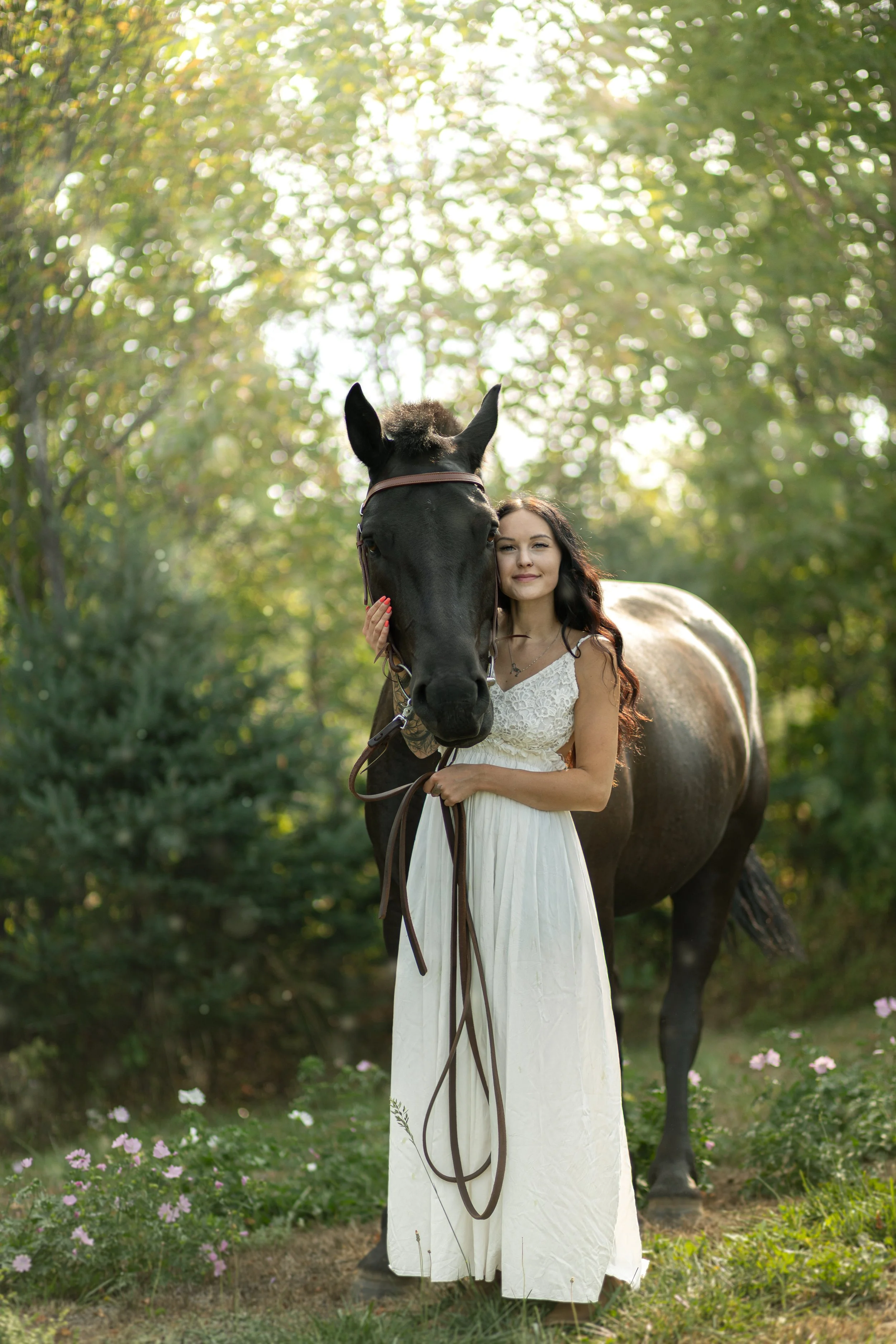 A woman in a white dress standing next to a black horse in a green outdoor setting during daylight.