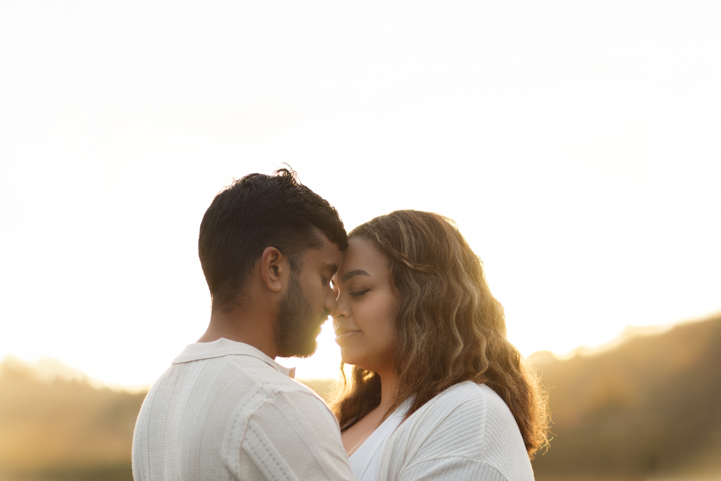 A man and woman with foreheads touching, eyes closed, outdoors during sunset, embracing each other.