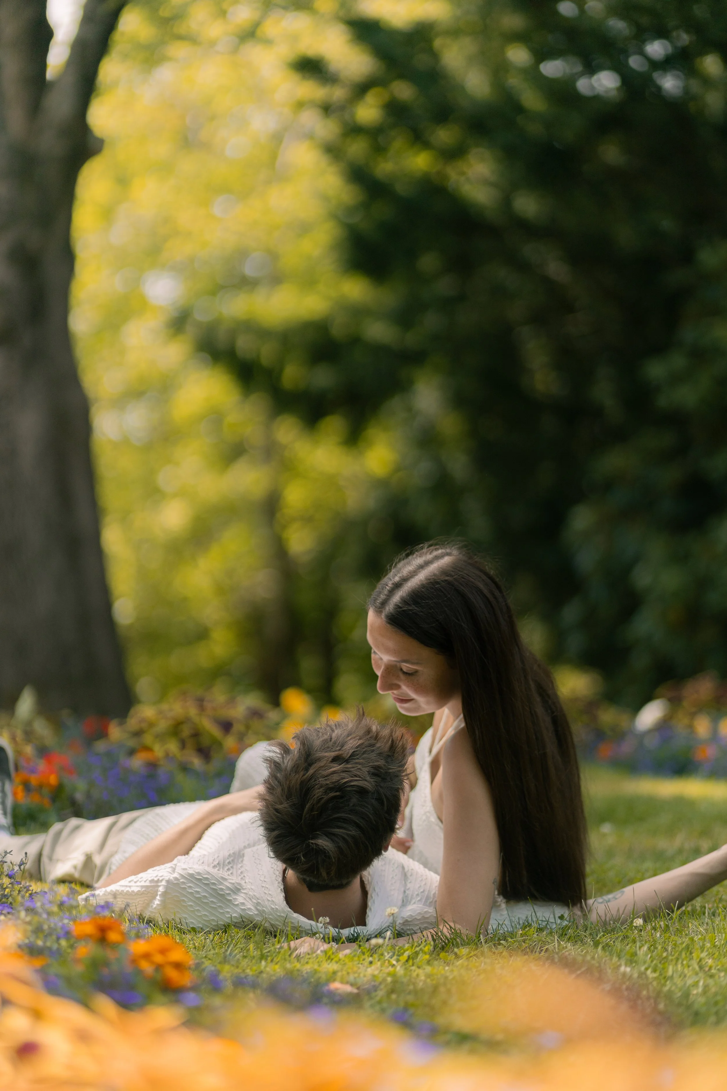 A woman and a child lying on grass in a park surrounded by flowers, with trees and sunlight in the background, enjoy a moment together.