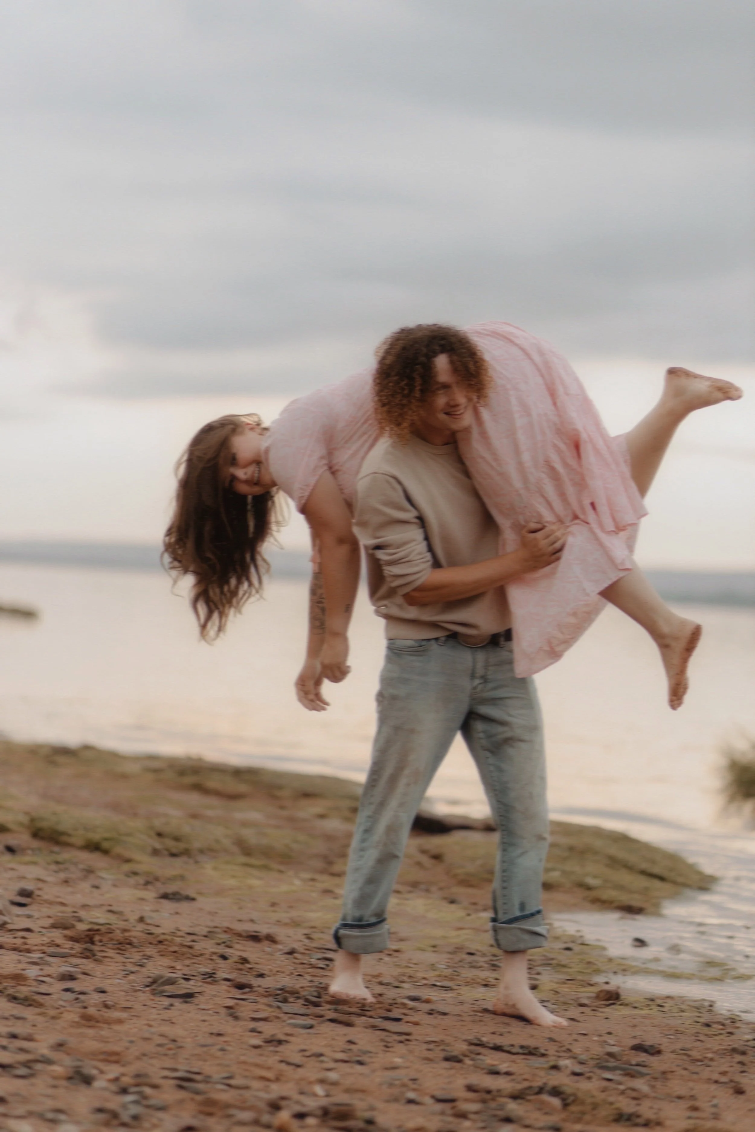 A man is carrying a woman on his back on a beach near water during cloudy weather.