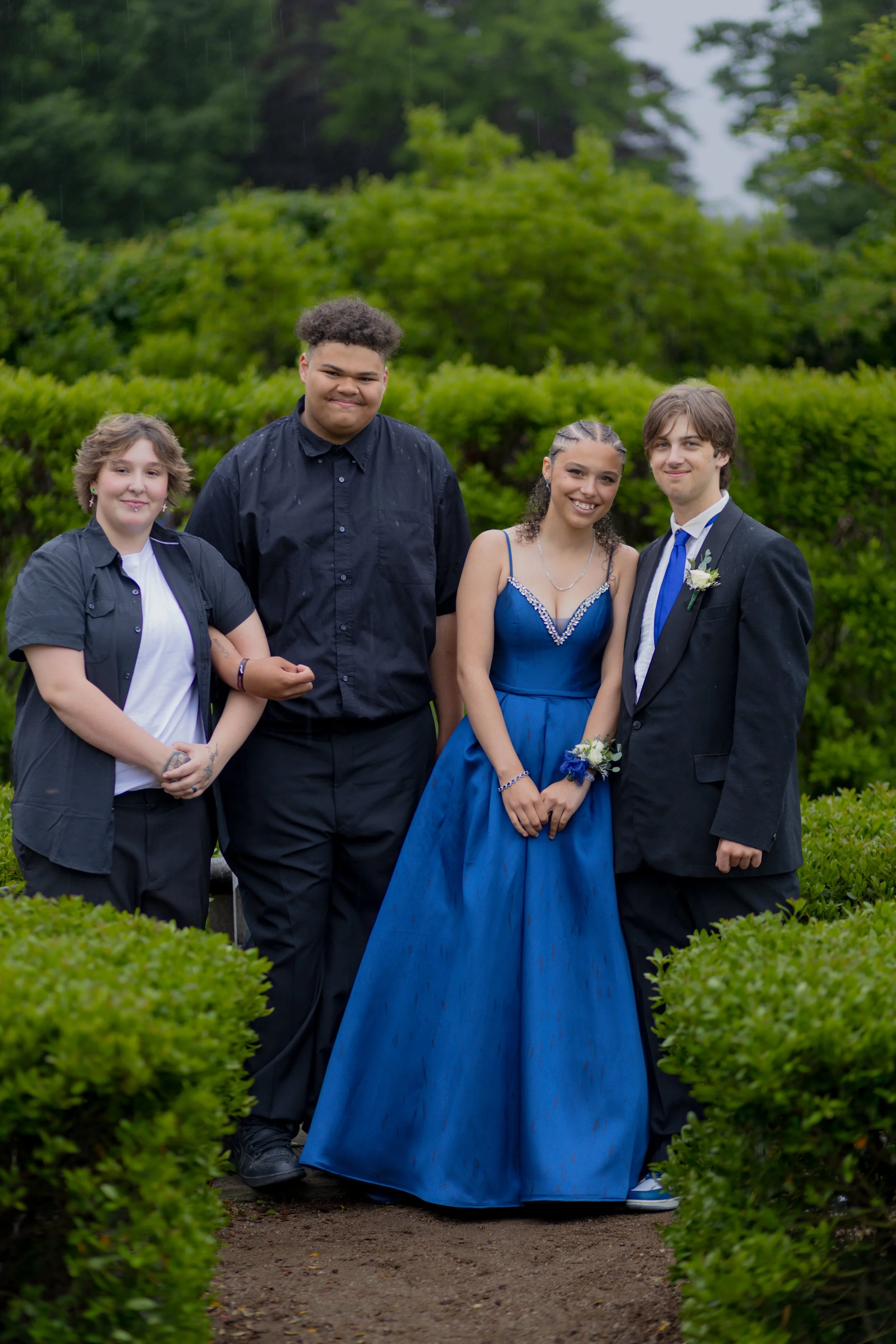 Four teenagers, two boys and two girls, standing outdoors with greenery in the background. One girl is wearing a formal blue dress, and the other girl is dressed casually. One boy is in a black suit with a boutonniere, and the other girl has tattoos 