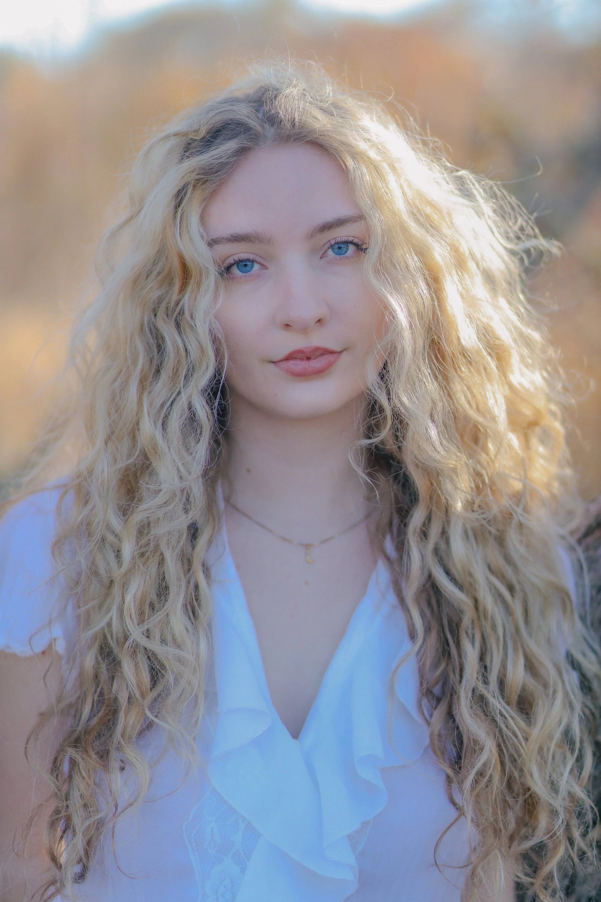 A young woman with long, curly blonde hair and blue eyes, wearing a white blouse with ruffles and a delicate gold necklace, standing outdoors with a blurred background of trees and sunlight.