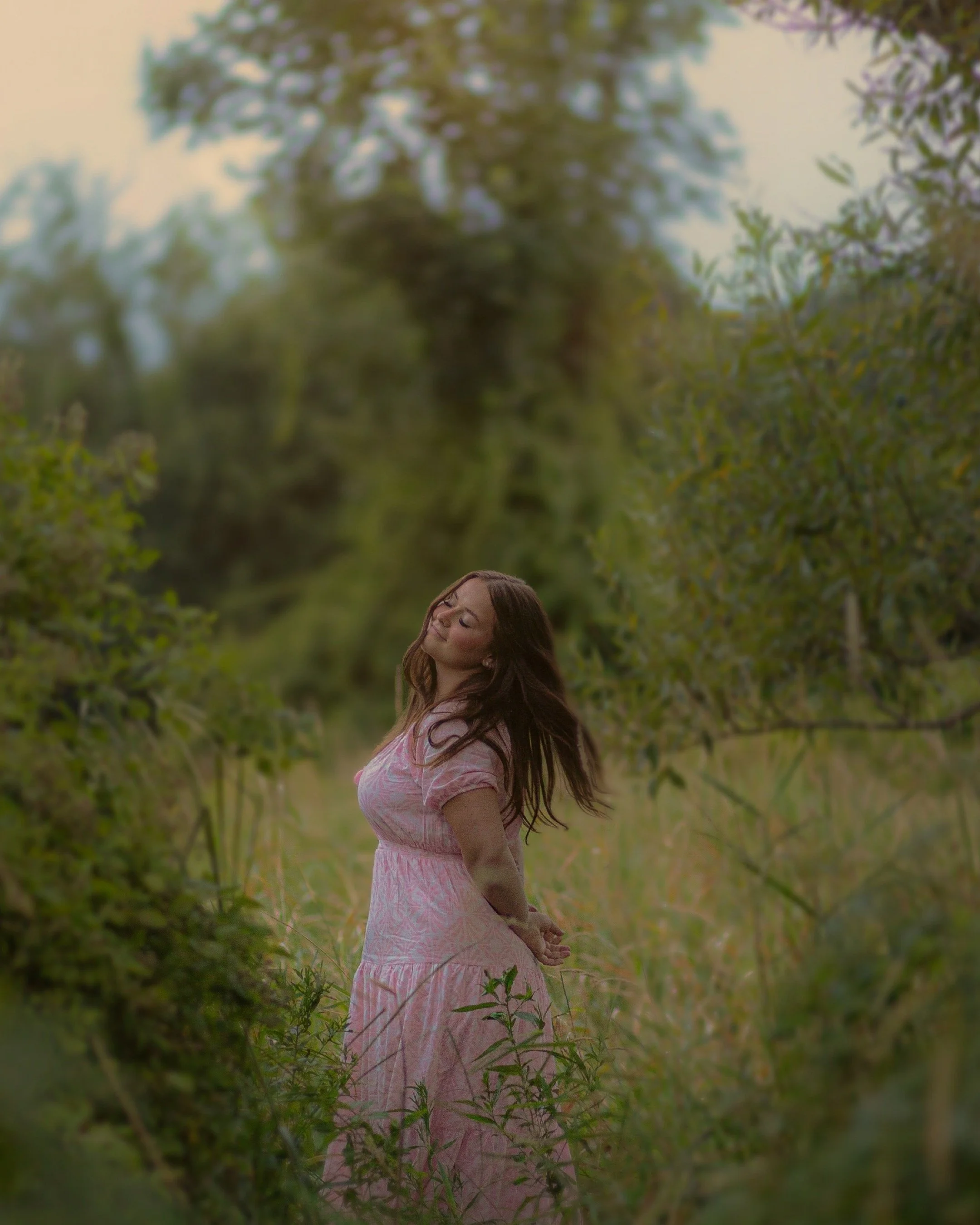 A woman in a pink dress standing among tall grass and trees with her eyes closed and arms behind her back, enjoying the outdoor setting.